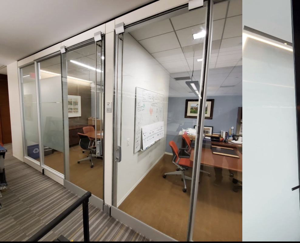 Glass-walled conference room with a desk, chairs, whiteboard, and framed artwork inside an office building.