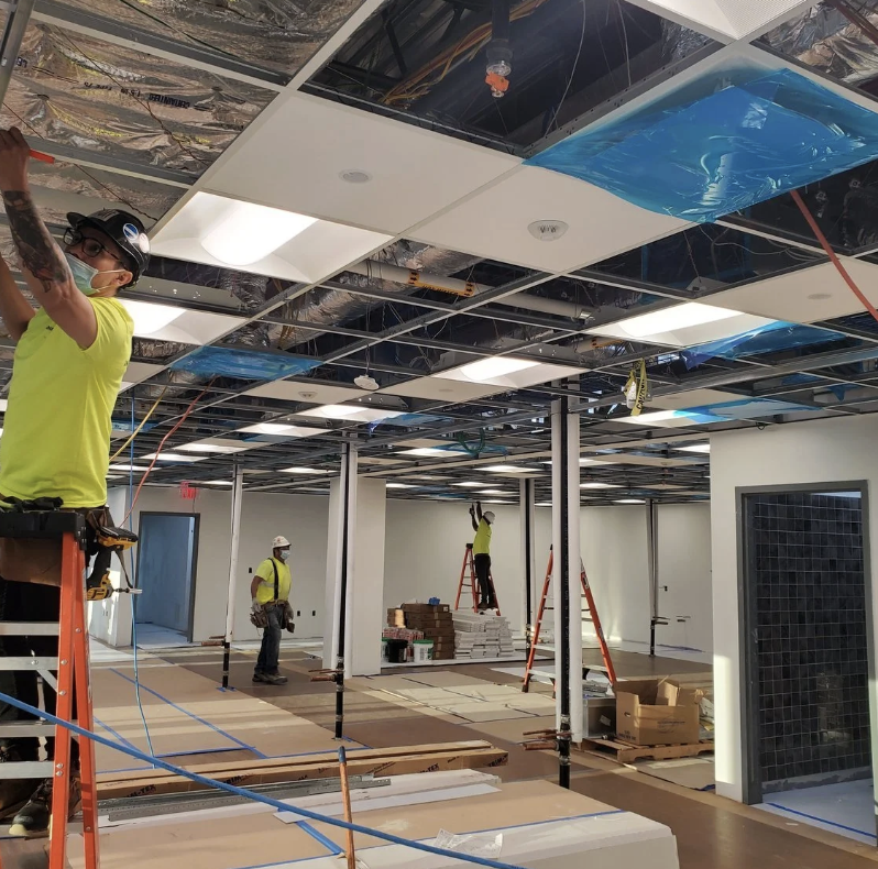 Construction workers installing ceiling tiles in an office space, with some workers on ladders and others on the floor, surrounded by building materials and tools.
