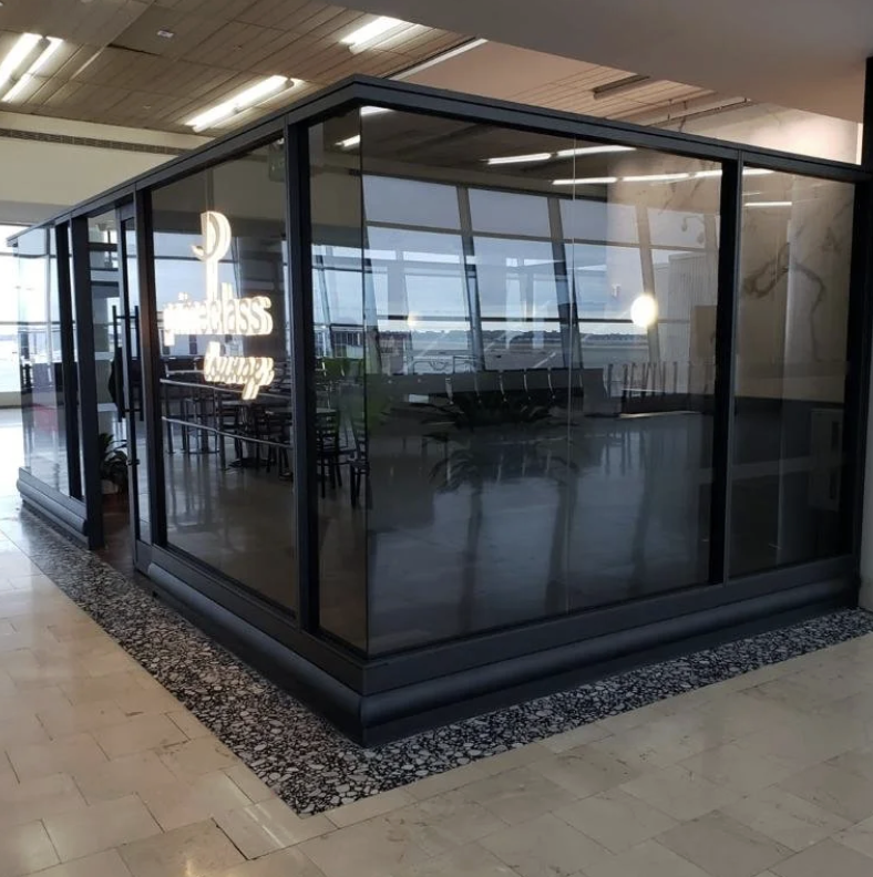A black-framed glass enclosure with sliding doors located in an indoor setting, possibly an airport or office. Inside the enclosure, there are chairs lined up against the windows. Reflection of ceiling lights and a neon sign reading 'first class loun