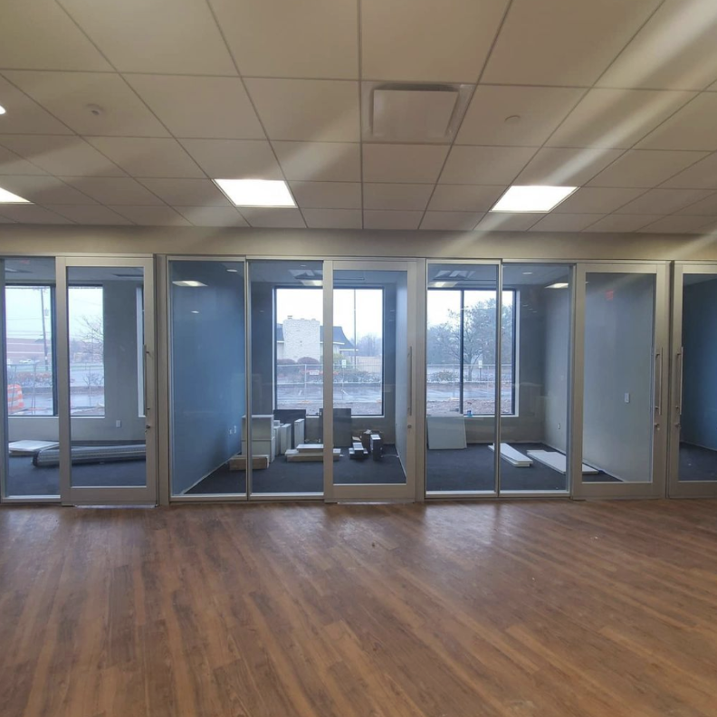Empty office space with glass walls and hardwood flooring. Some construction materials are inside the enclosed area, indicating ongoing construction or renovation.