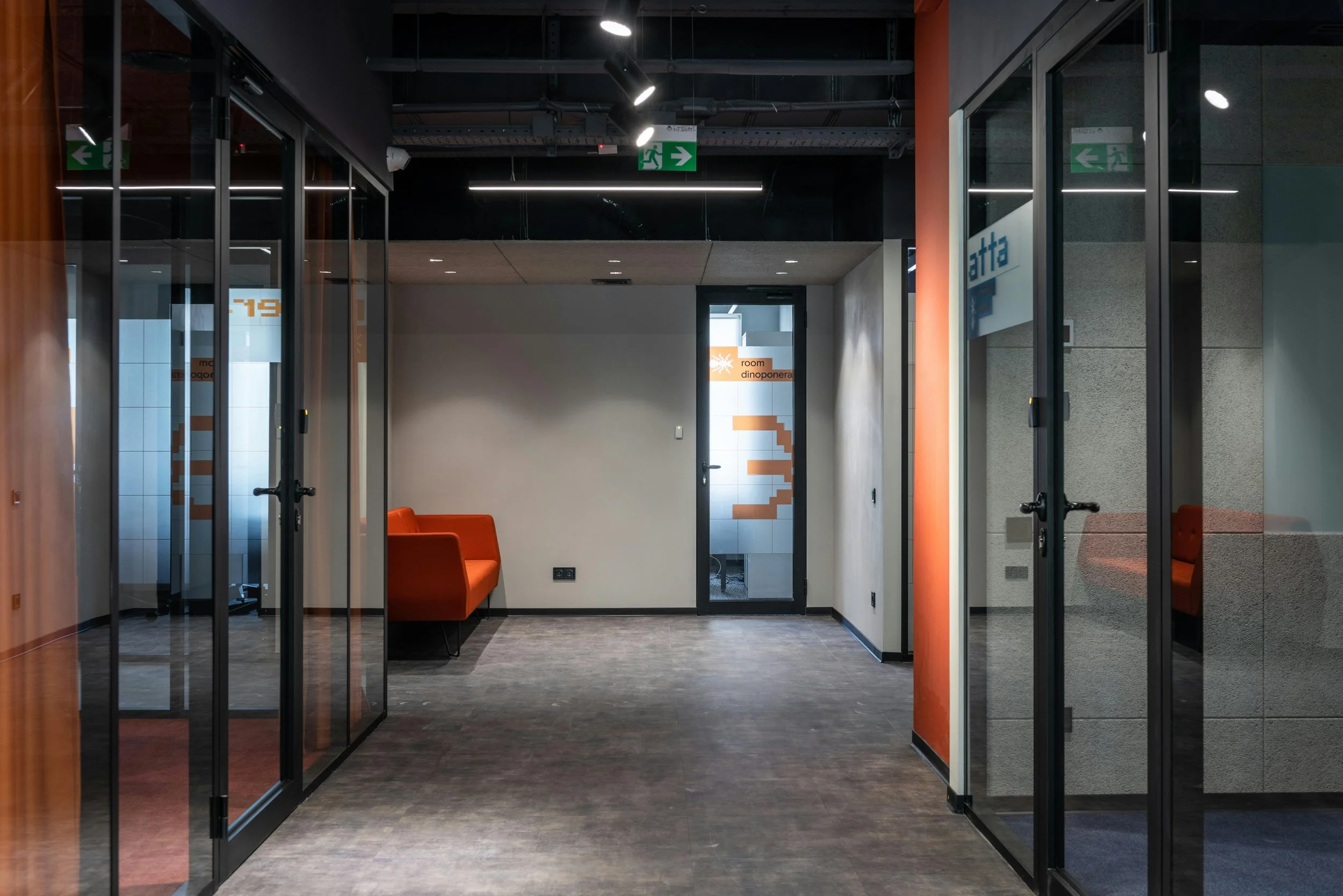 Modern office corridor with glass doors, orange accents, and a small seating area with an orange sofa.