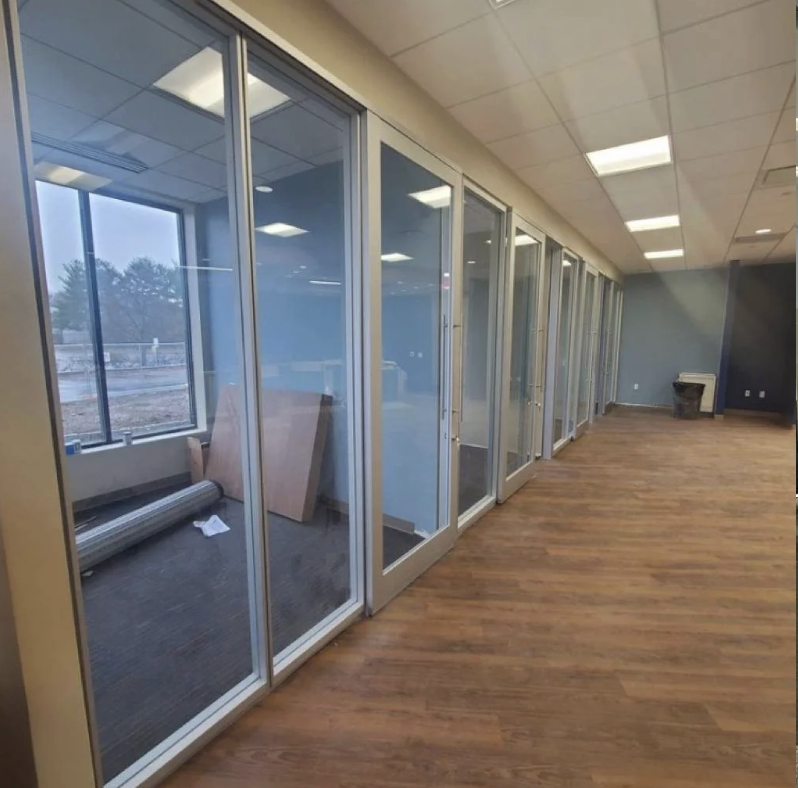 Empty office space with glass partition walls, wooden flooring, and construction materials inside one room.