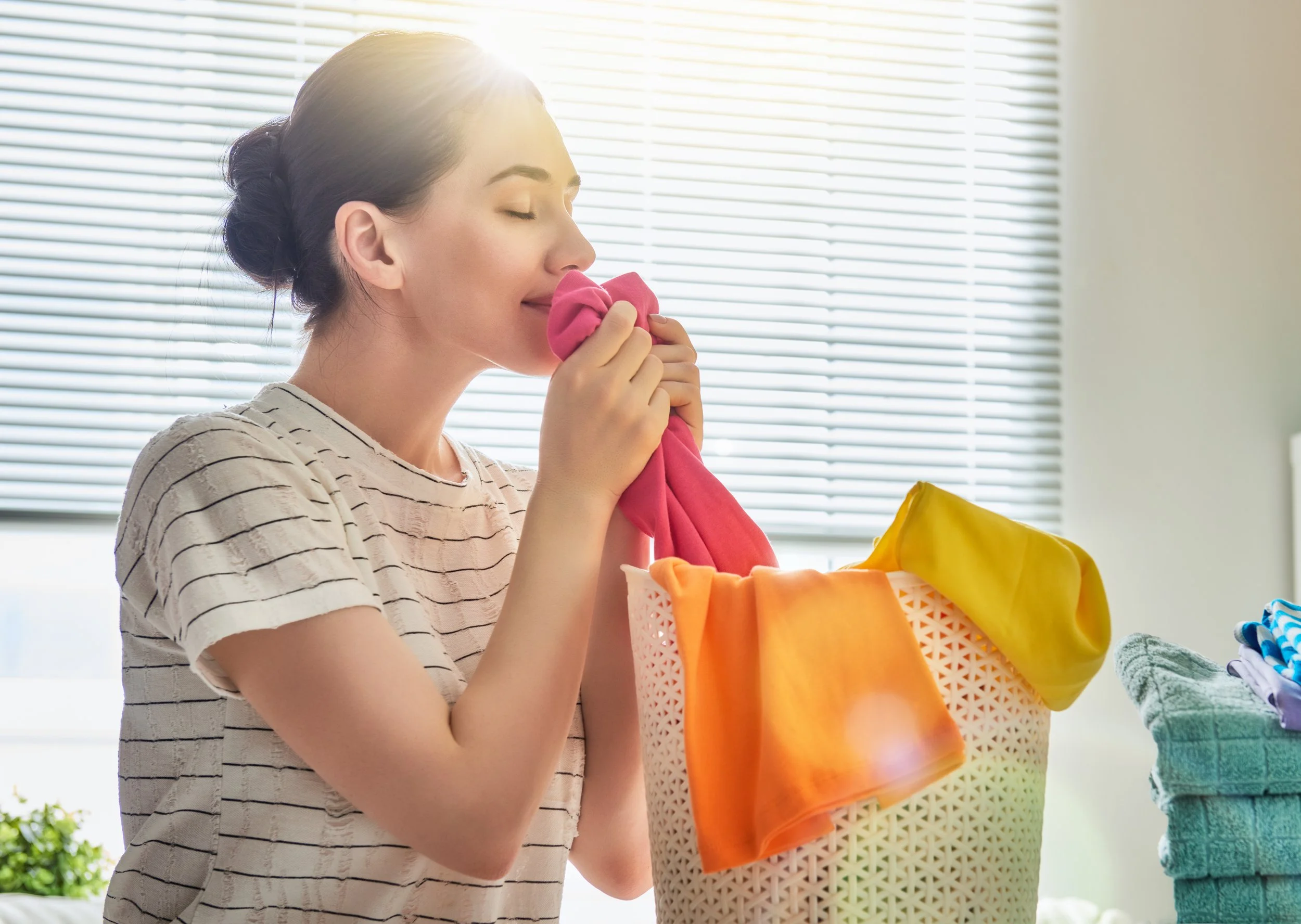 A woman with dark hair in a bun, wearing a striped shirt, smells a pink cloth while doing laundry in a bright room with sunlight and window blinds.