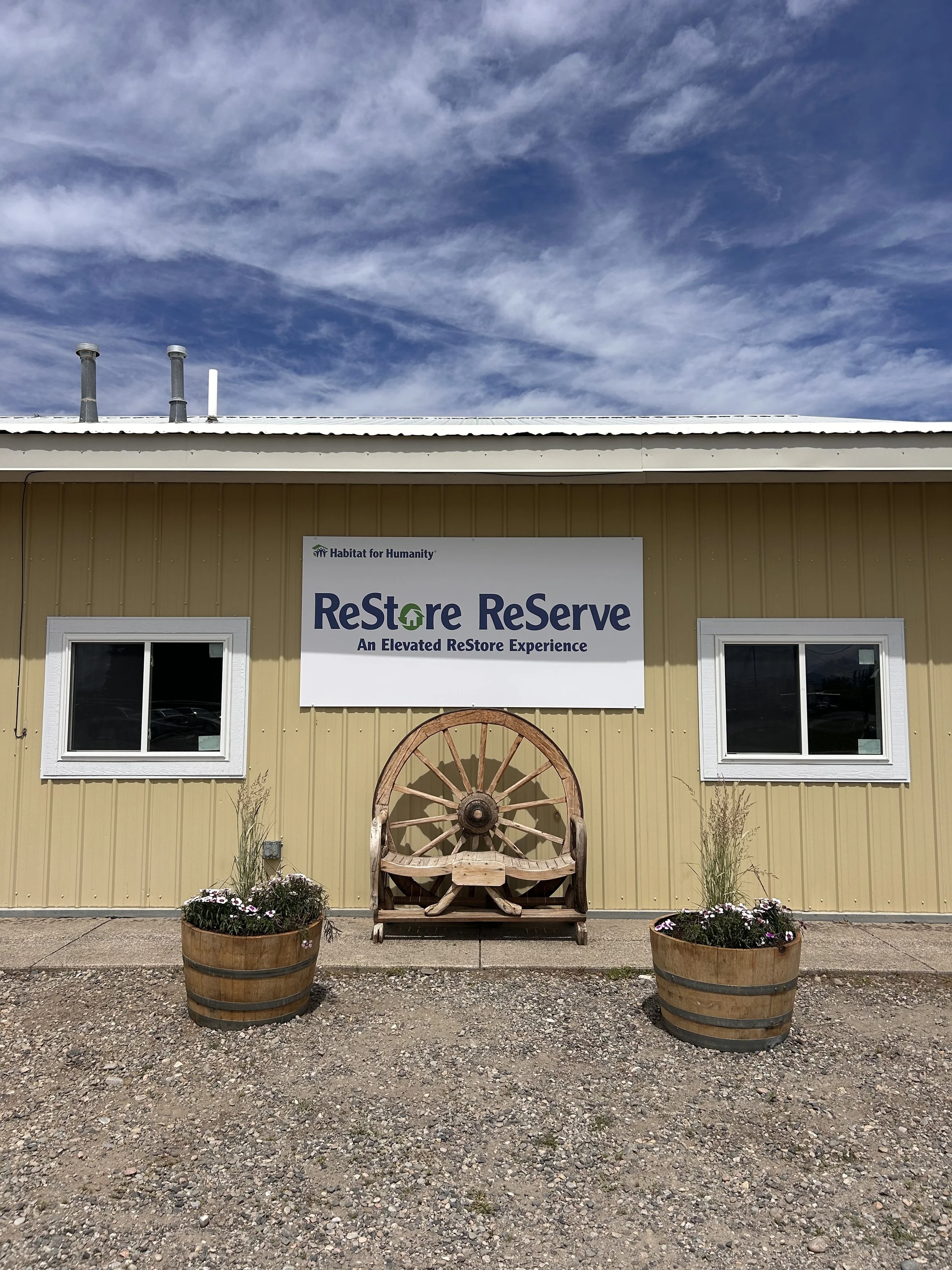 Outside, front of the ReStore ReServe focused on the sign reading "Habitat for Humanity of Gallatin Valley ReStore ReServe. An elevated ReStore experience." Features a bench and two flower pots in front.