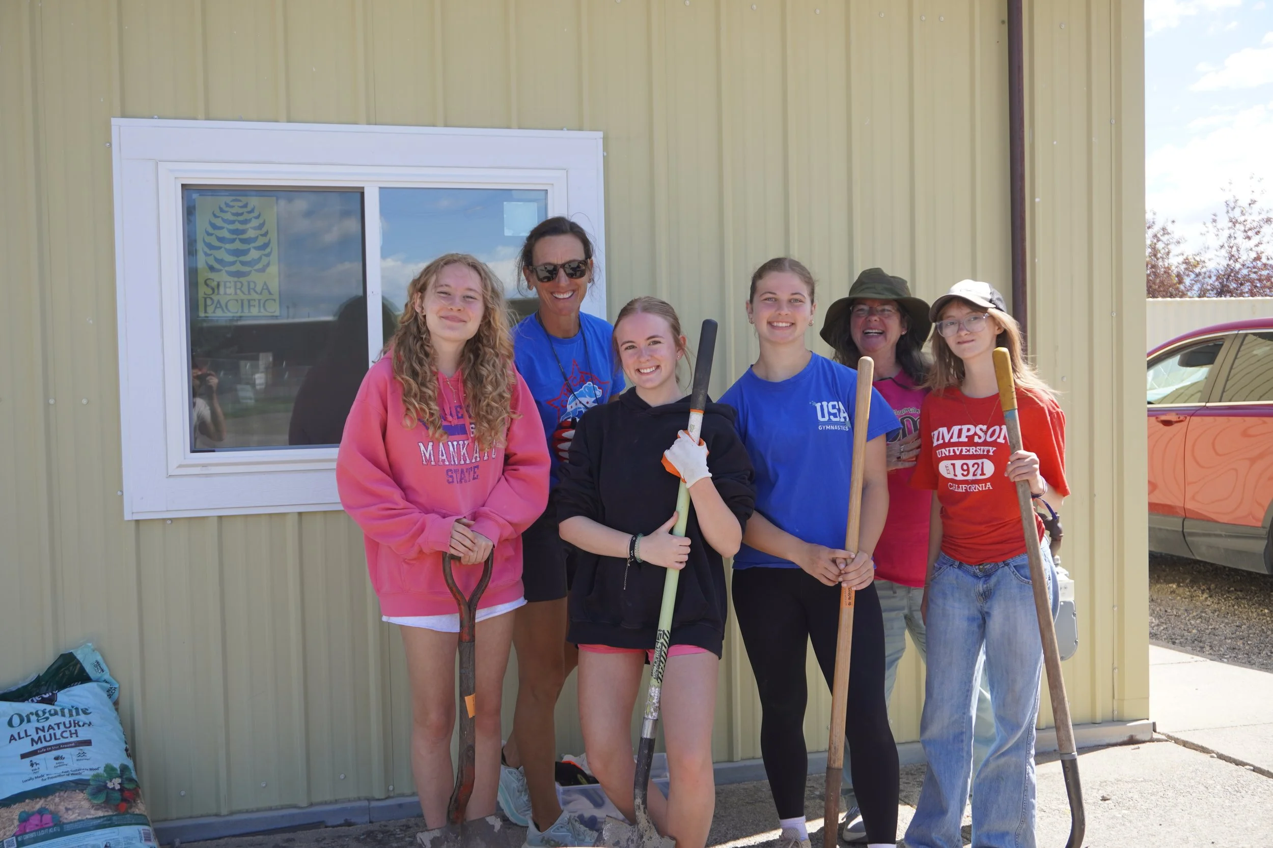 A group of four young women and two older women standing in front of a yellow building with a window, holding gardening tools, smiling at the camera.