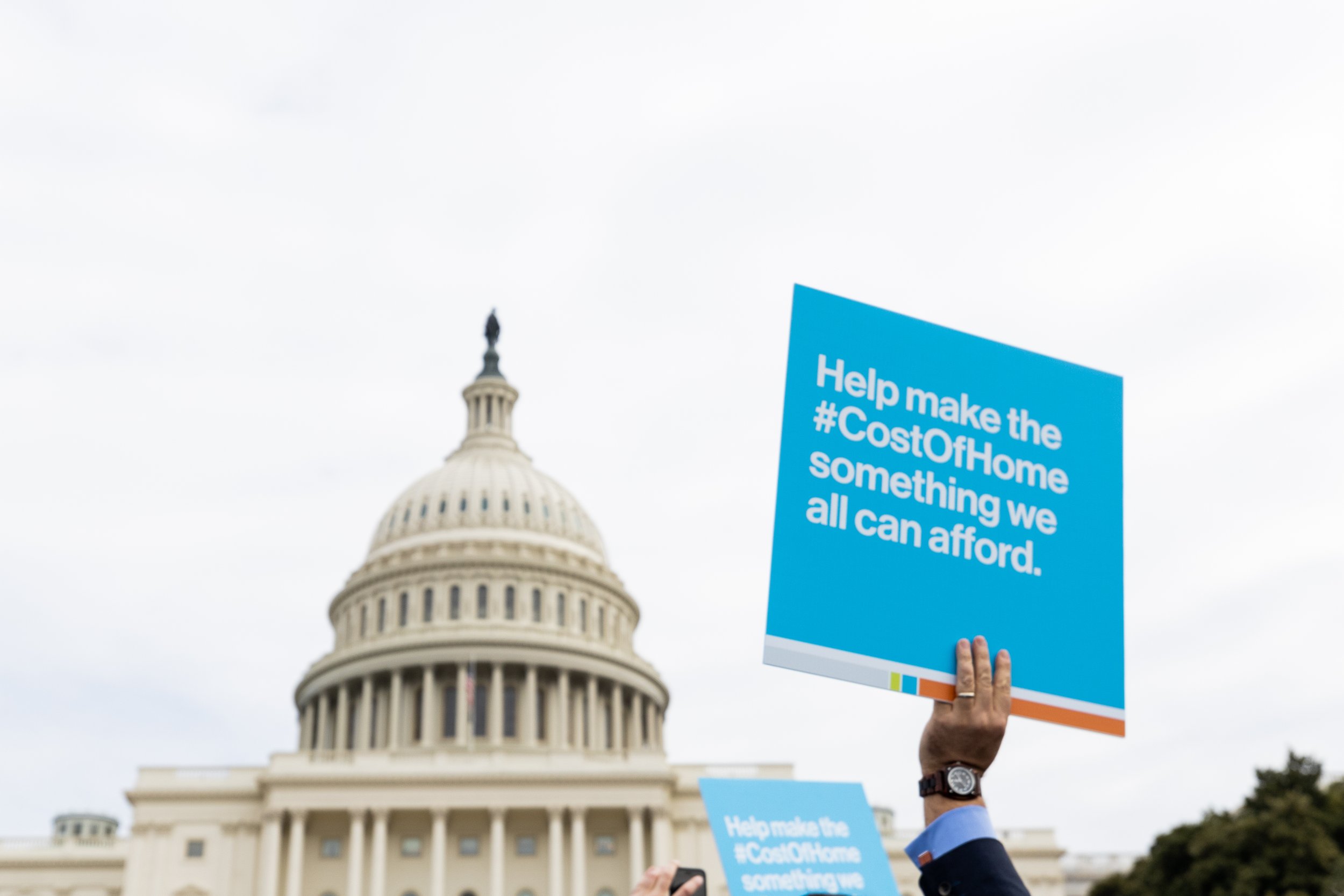 Person holding a blue protest sign in front of the U.S. Capitol building. The sign reads: 'Help make the #CostOfHome something we all can afford.' The sky is overcast.