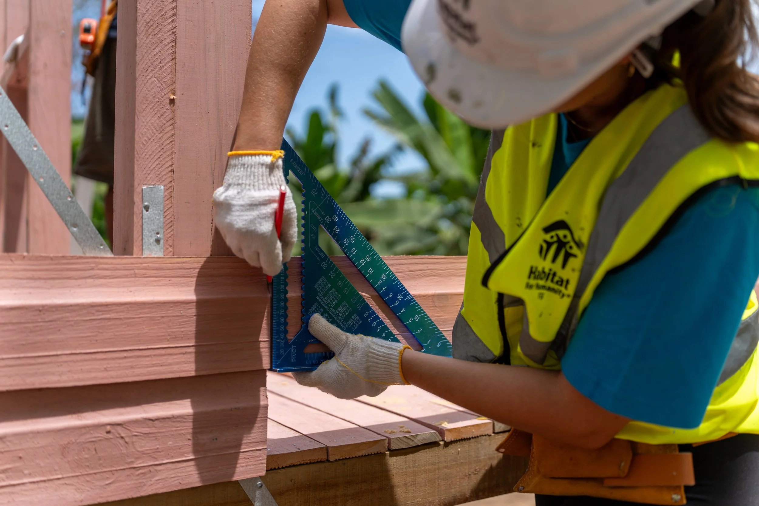 A person wearing a safety vest and gloves using a carpenter's square to measure and mark a wooden structure during construction.