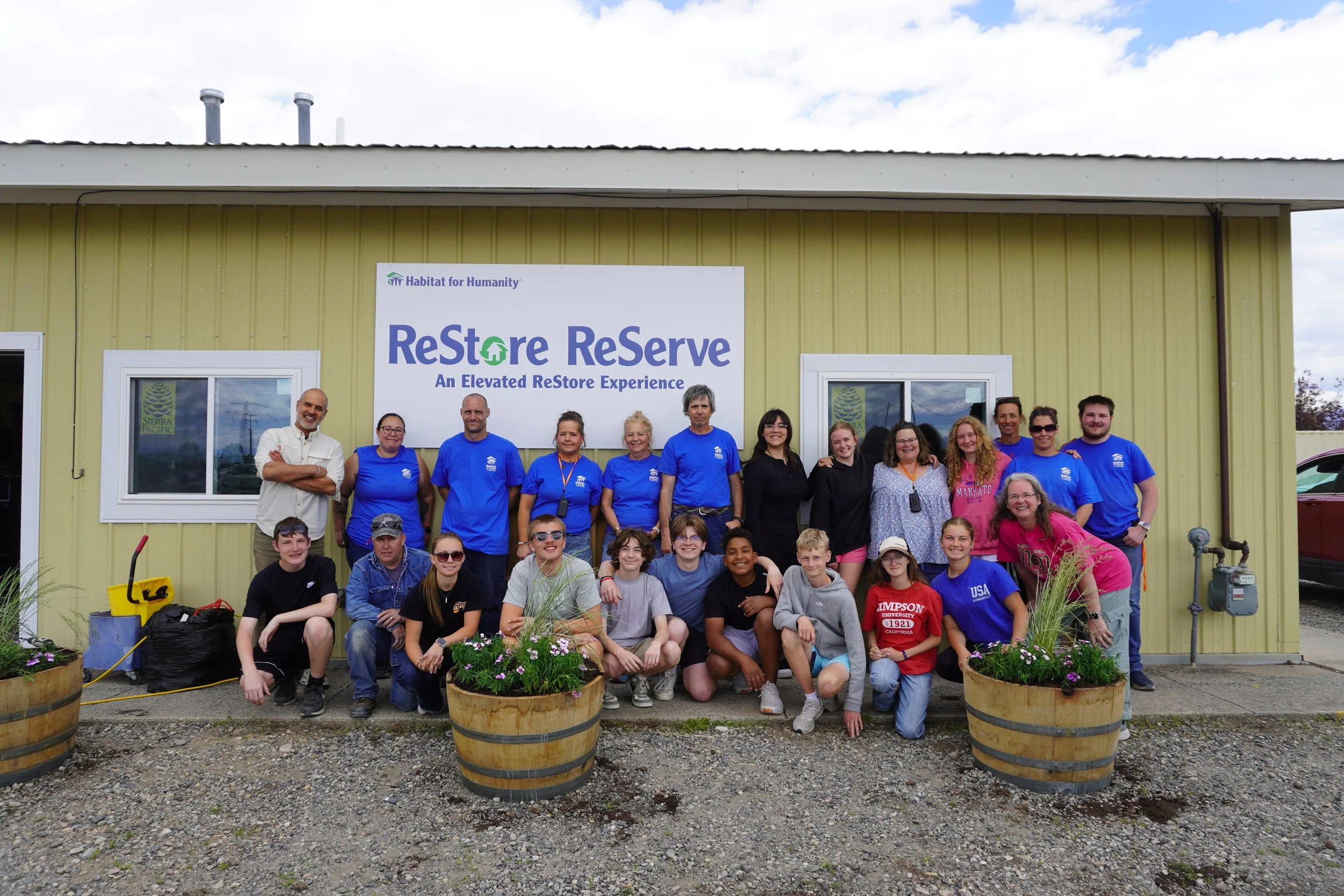 Group of people standing and sitting in front of a building with a Habitat for Humanity sign that reads 'Restore Reserve: An Elevated ReStore Experience.' Four large flower pots with pink and purple flowers are in the foreground.