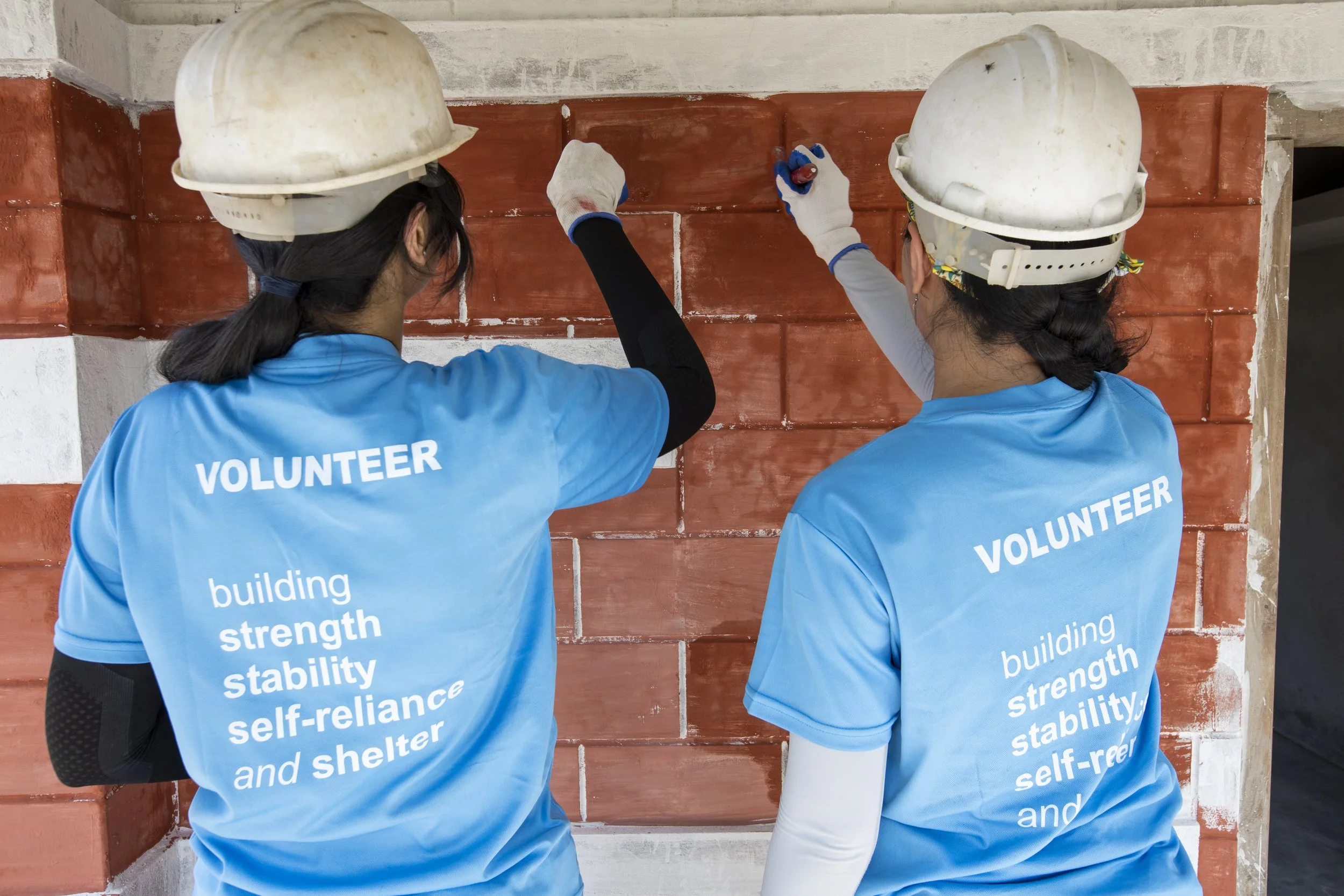Two volunteers wearing blue shirts and white construction helmets working on a brick wall. The shirts have words related to building strength, stability, and self-reliance. One volunteer is using a trowel to apply mortar or cement onto the bricks.