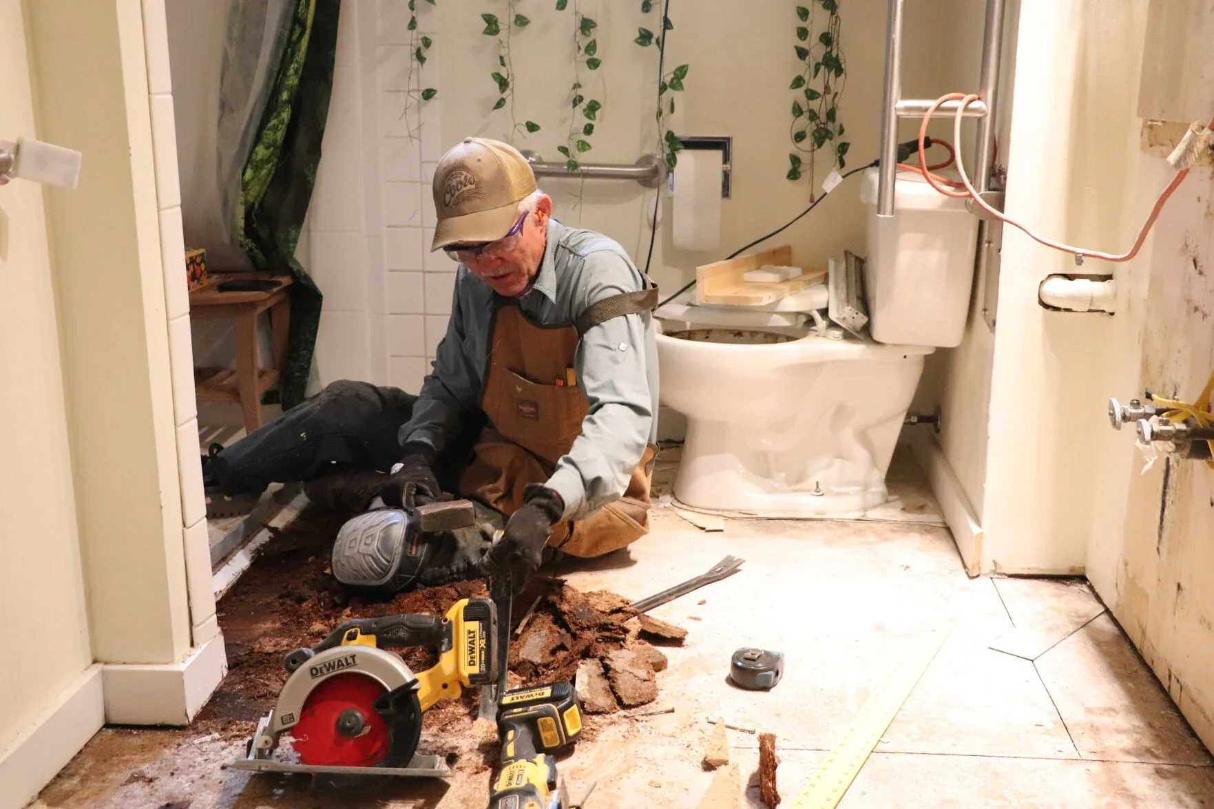 A man working on bathroom plumbing repair, sitting on the floor with tools around him, near a toilet and partially removed floor tiles.