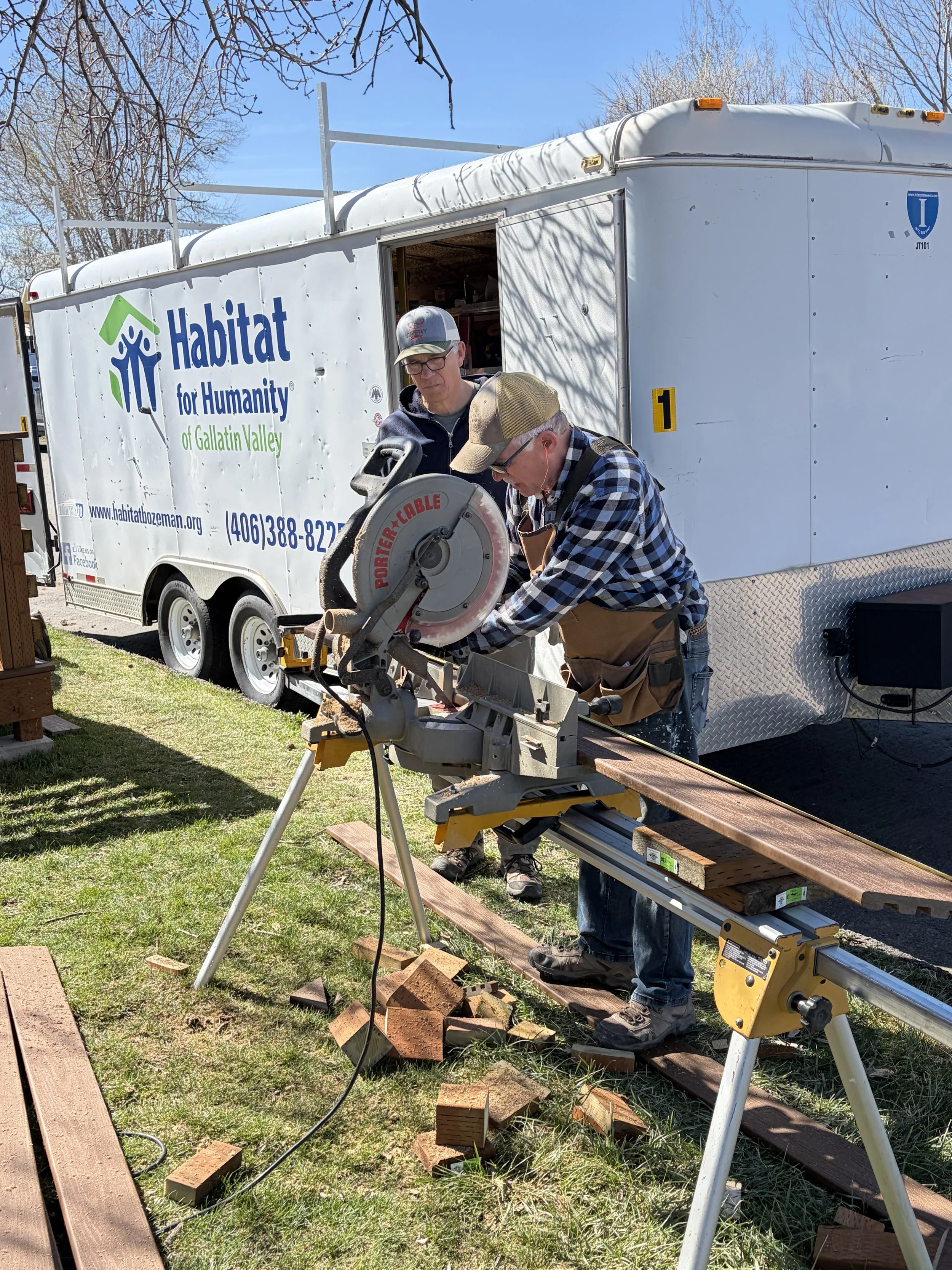 Two men working outdoors with a portable saw, cutting wood or bricks, next to a white Habitat for Humanity trailer, with wood planks on the ground, trees, and a clear sky in the background.