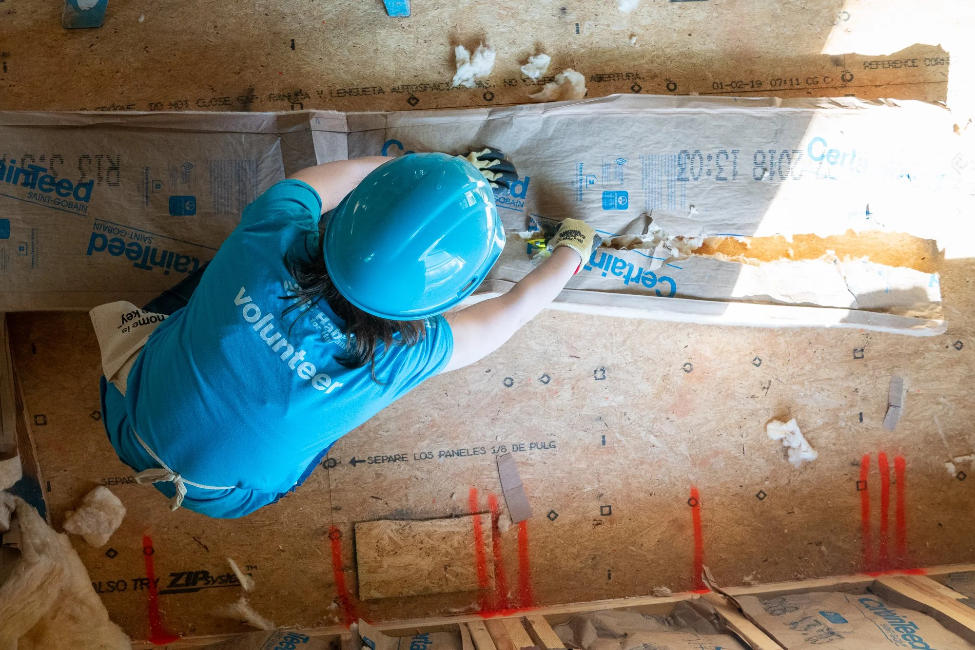 Worker in a blue shirt, yellow gloves, and a blue safety helmet installing insulation in a wall cavity with wooden framing, insulation rolls, and construction materials visible.