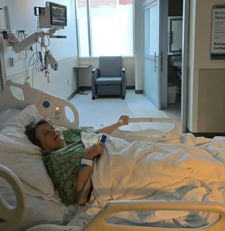 A patient lying in a hospital bed in a hospital room, with medical monitoring equipment attached, and an arm extended outside the bed holding a long medical tube.
