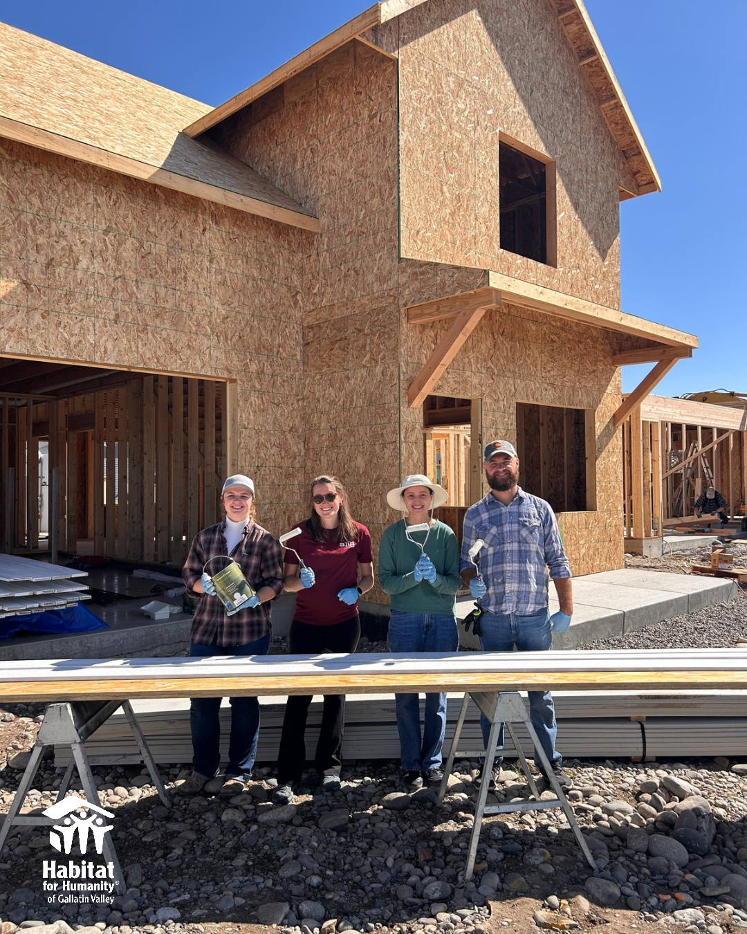 Four volunteers standing in front of a partially constructed house, holding paint rollers and paint cans, smiling at the camera. The house is made of wood and OSB sheathing, and the sky is clear and blue. Habitat for Humanity logo is visible in the lower left corner.