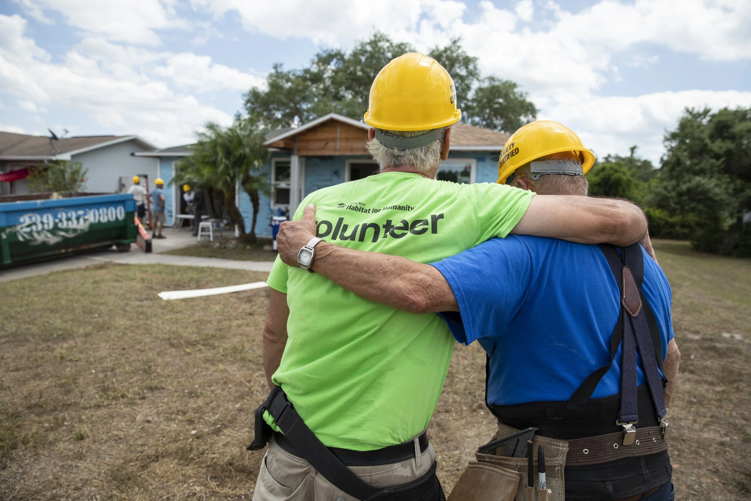 Two male volunteers wearing hard hats with their arms around each other looking at the house being built.