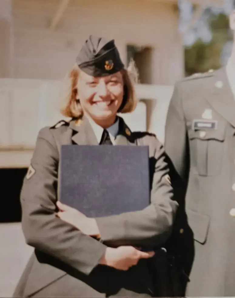 A woman in a military uniform smiling and holding a dark folder or document, standing next to a man also in military uniform.