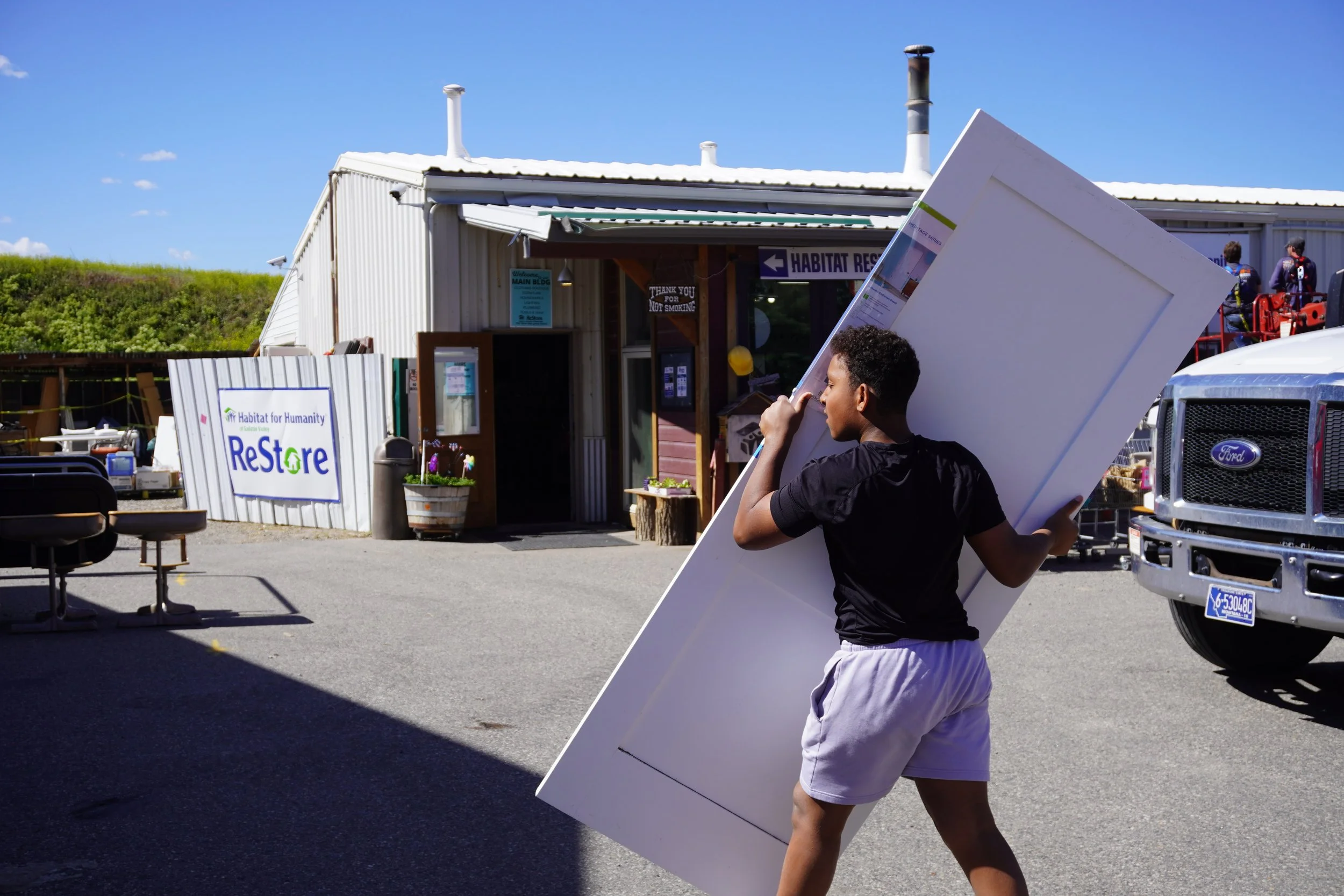 A young man carrying a large white panel during a Habitat for Humanity event in a parking lot outside a building with a sign for Habitat ReStore.