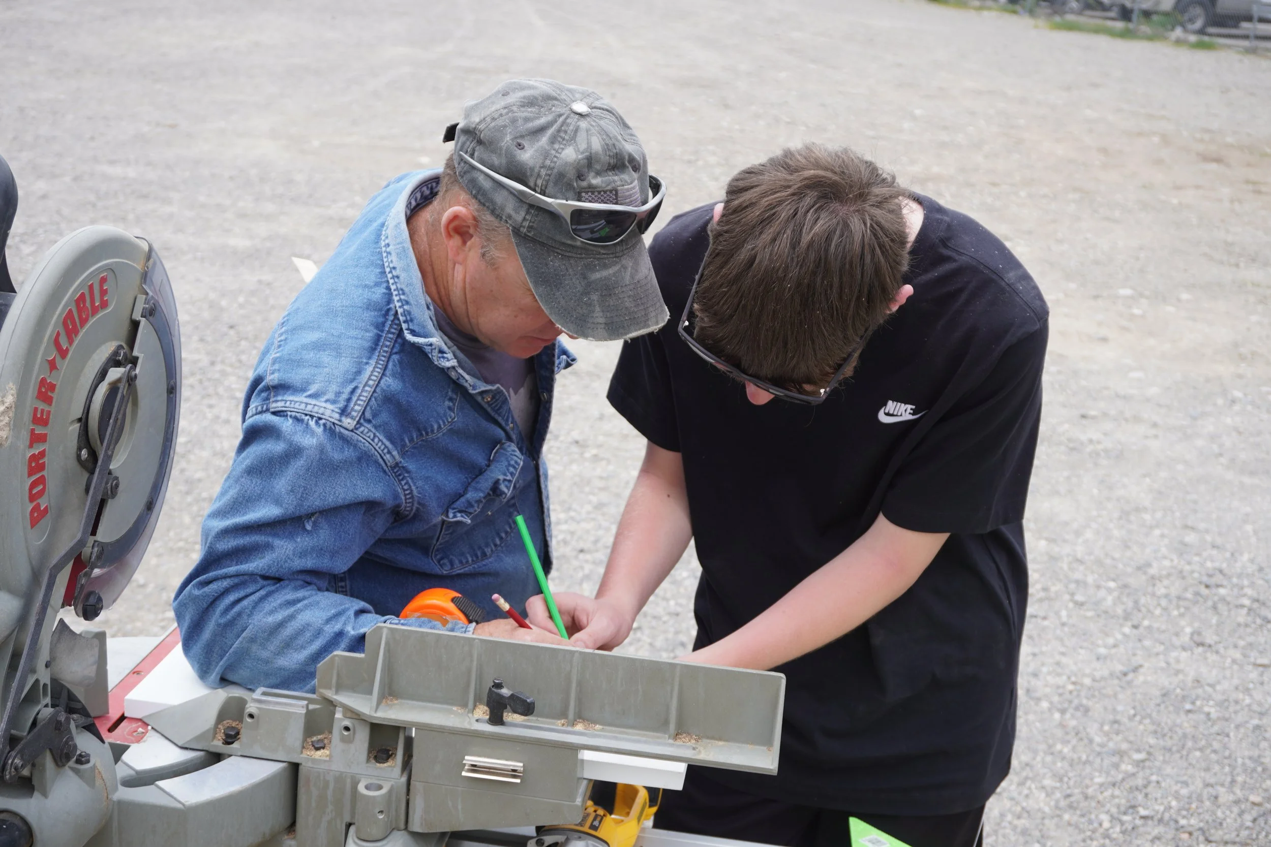 Two men working together outdoors beside a large saw, with one man wearing a denim jacket and baseball cap and the other a black Nike t-shirt and glasses.