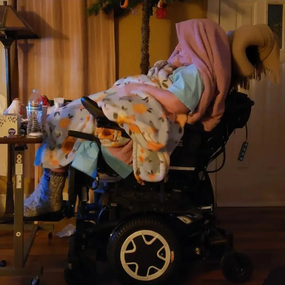 An elderly person sitting in a motorized wheelchair, covered with a floral blanket, with a pink towel over their head, in a cozy indoor setting.