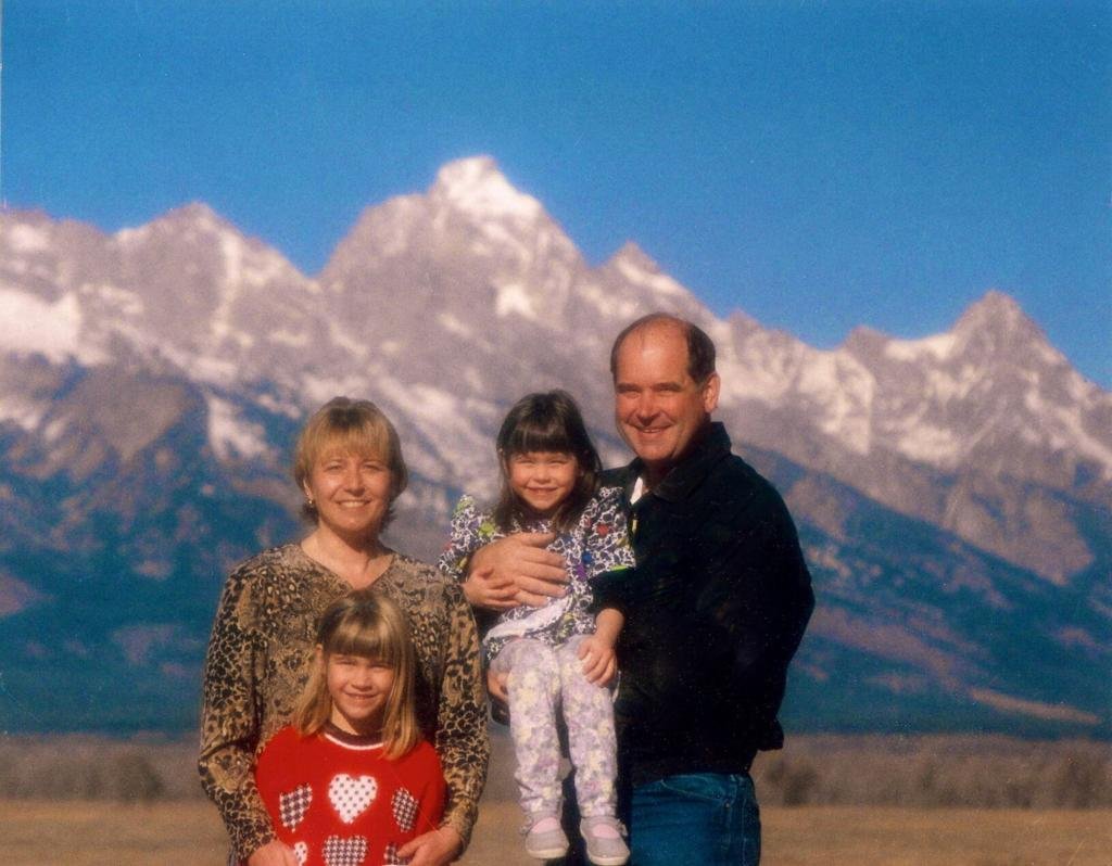 A family of four posing outdoors with snow-capped mountains in the background.