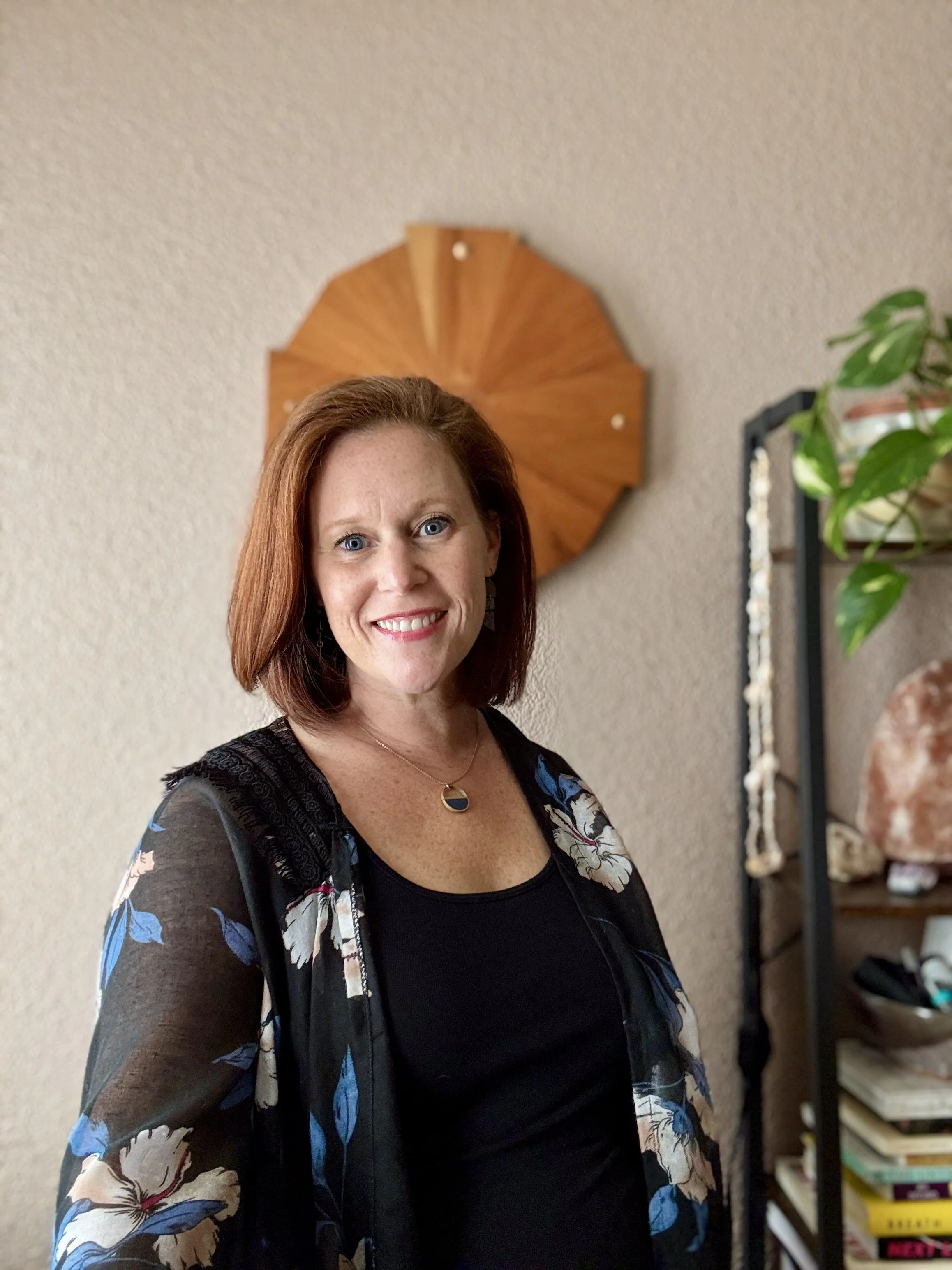 A smiling woman with red hair wearing a black top and a floral shawl, standing indoors in front of a beige wall with a wooden decorative piece and a shelving unit with books and decorative items.