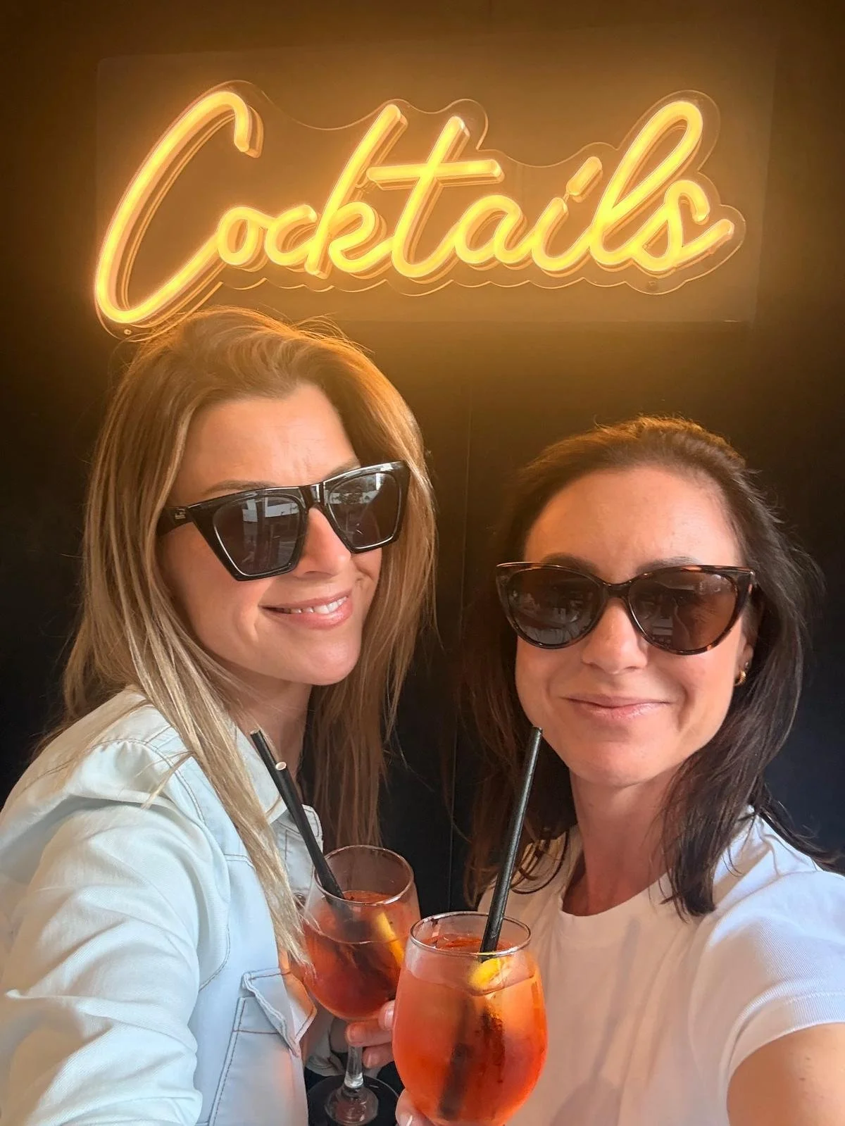 Two women wearing sunglasses and smiling, holding cocktails with straws and lemon slices, standing in front of a black wall with a yellow neon sign that says 'Cocktails'.