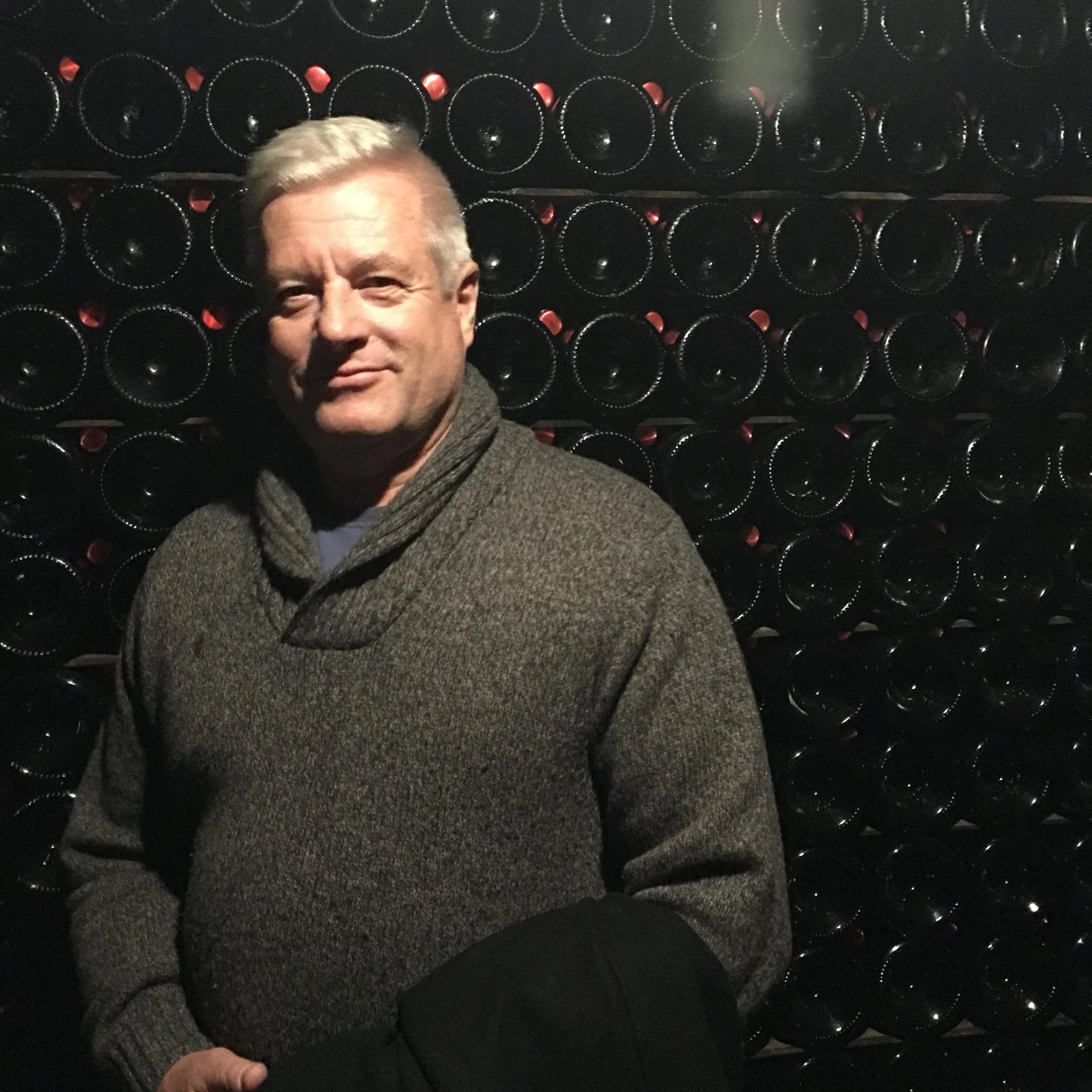 A man with light-colored hair wearing a gray sweater, standing in front of a wall of wine bottles.
