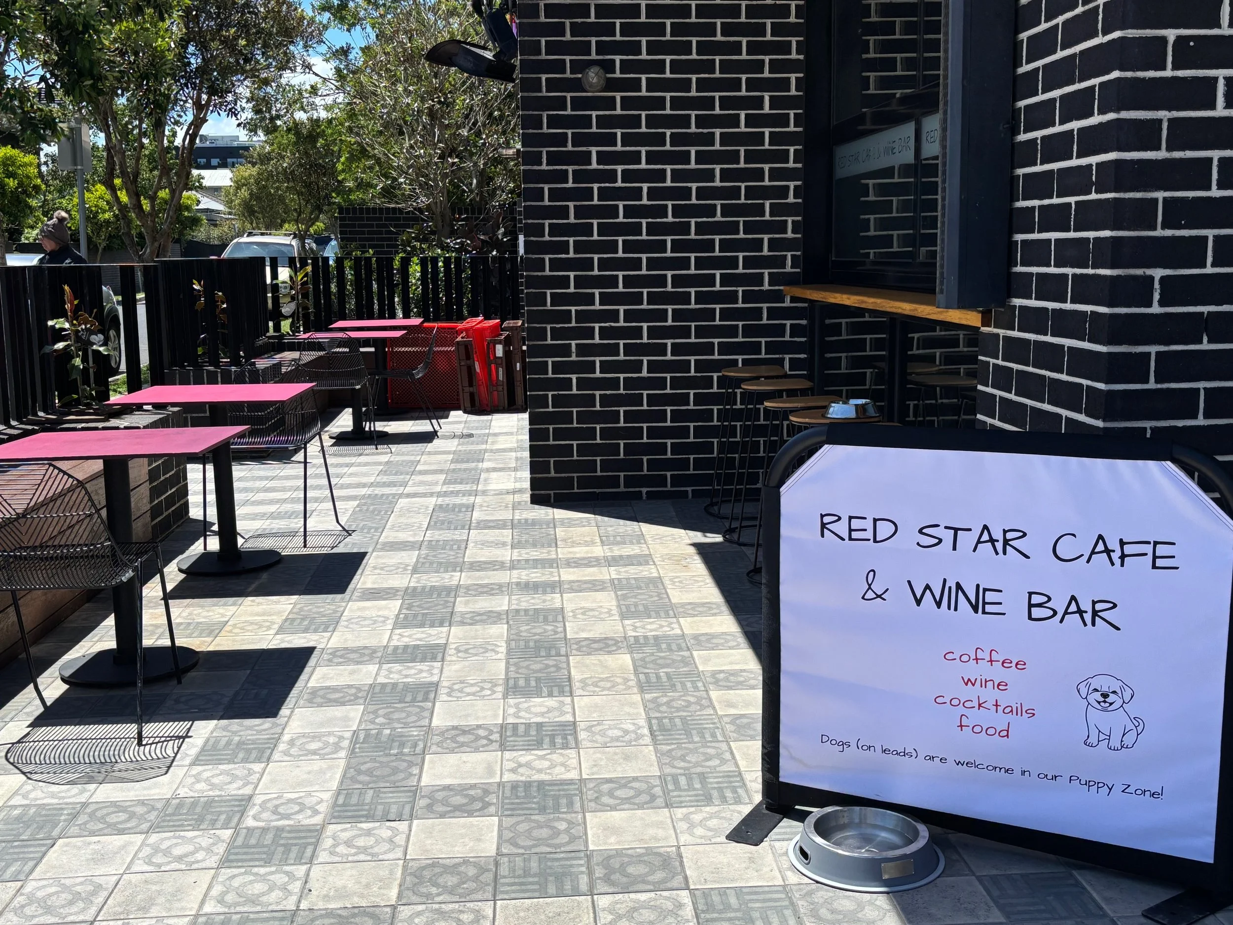 Outdoor patio at Red Star Cafe & Wine Bar with pink tables and black chairs, some red containers, and a signboard advertising coffee, wine, cocktails, food, and a puppy zone, with trees and parked cars in the background.