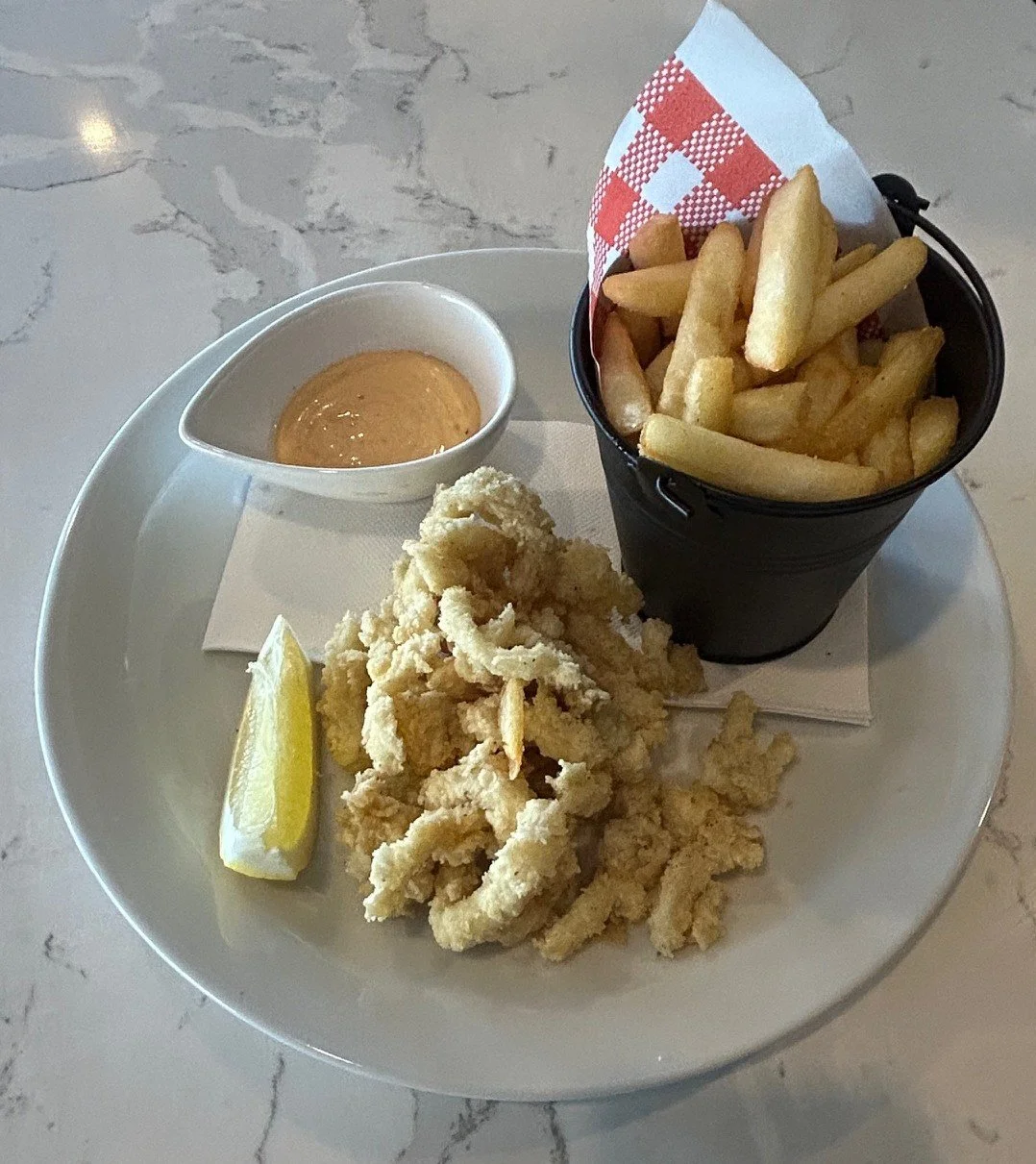 Fried calamari with lemon wedge, dipping sauce in a small white dish, and French fries in a black tin cup with checkered paper, on a white plate.