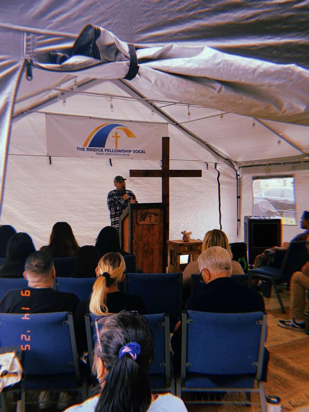 A man speaking at a podium with a Christian cross in a tent during a church or community gathering. Audience members are seated, and a banner reads 'The Bridge Fellowship SoCal'.