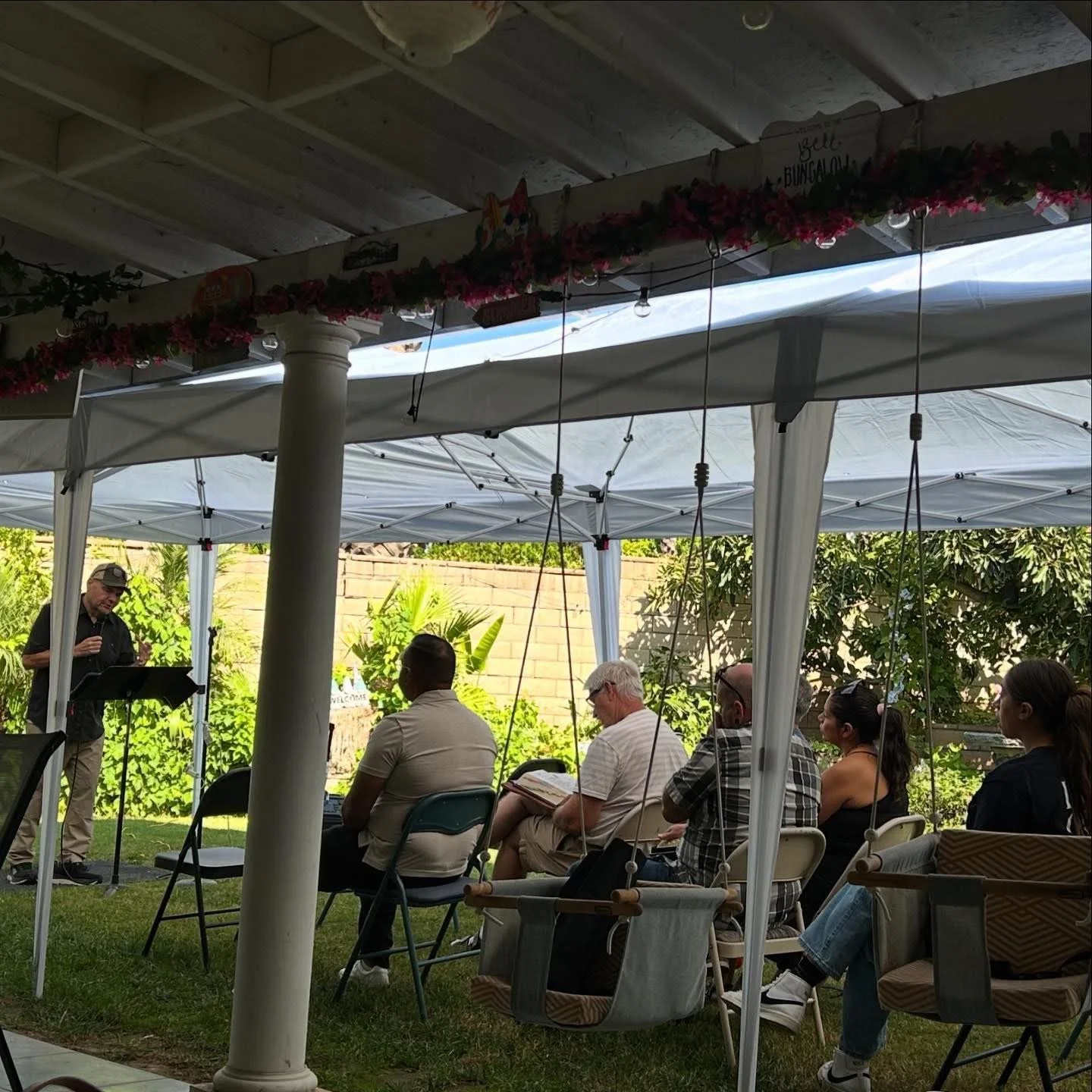 A man giving a presentation outdoors under a white canopy tent, with an audience seated in chairs and some in loungers, surrounded by greenery and a brick wall.