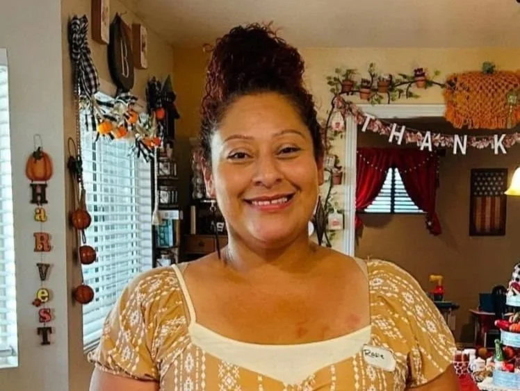 Smiling woman with curly hair styled in a bun, wearing a yellow patterned top, in a decorated room with Thanksgiving decorations including a "GIVE THANKS" banner and fall-themed ornaments.