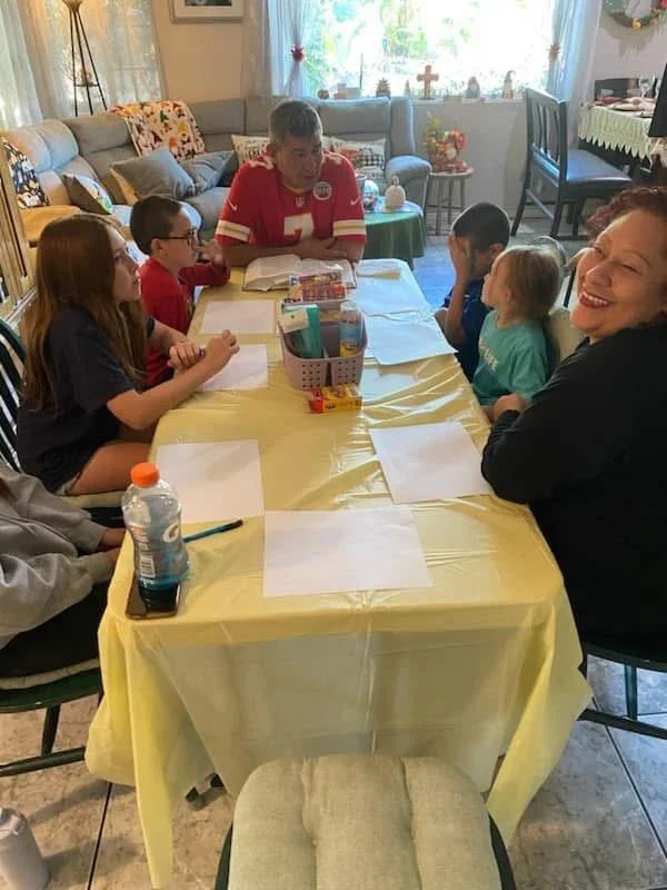 People gathered around a table with a yellow tablecloth, engaged in a discussion or activity, in a cozy living room with sunlight, plants, and holiday decorations in the background.