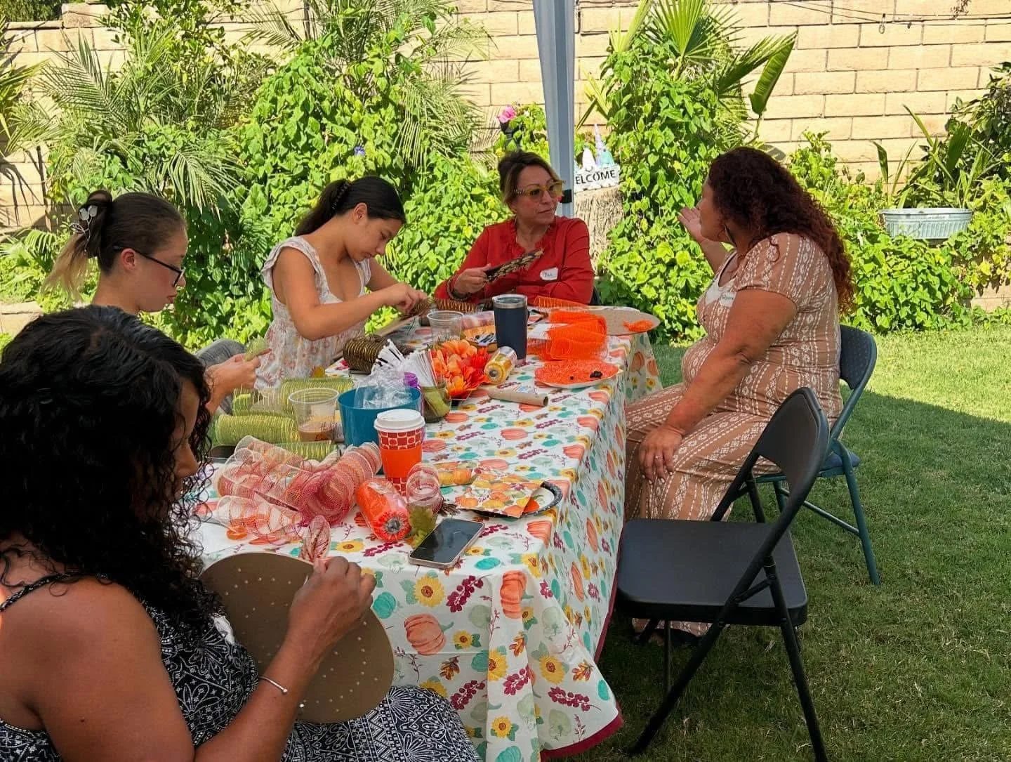 People sitting around a table outdoors engaging in a craft activity, with decorations and supplies on the table and greenery in the background.