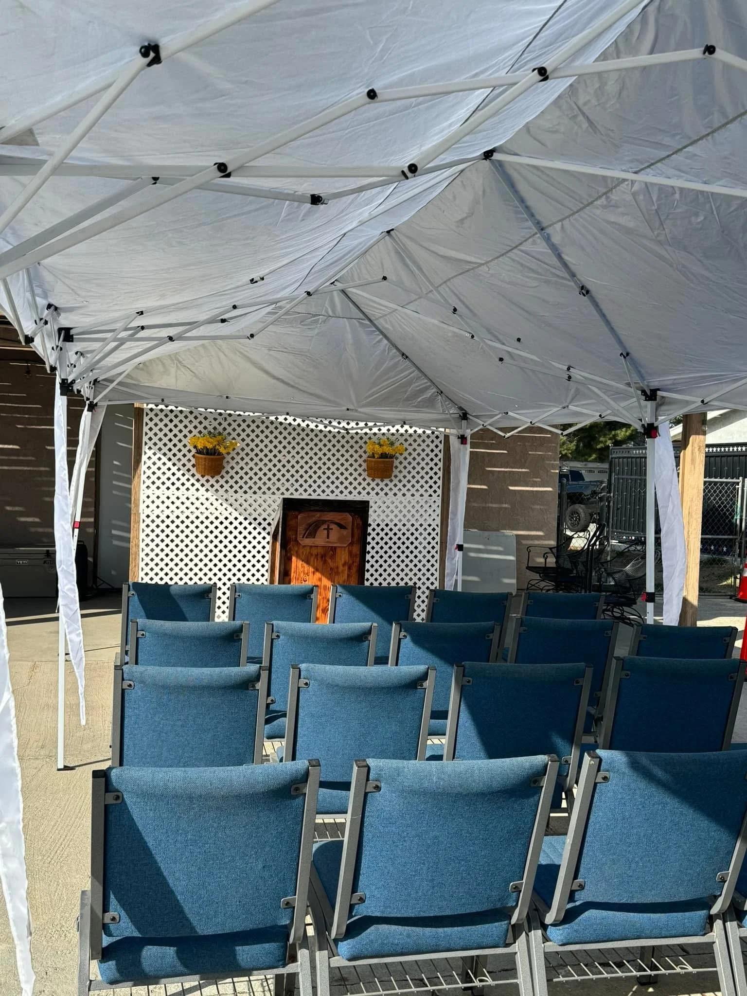 Outdoor seating area with blue chairs underneath a white canopy, facing a small wooden podium and a white lattice wall with two yellow flower baskets above.