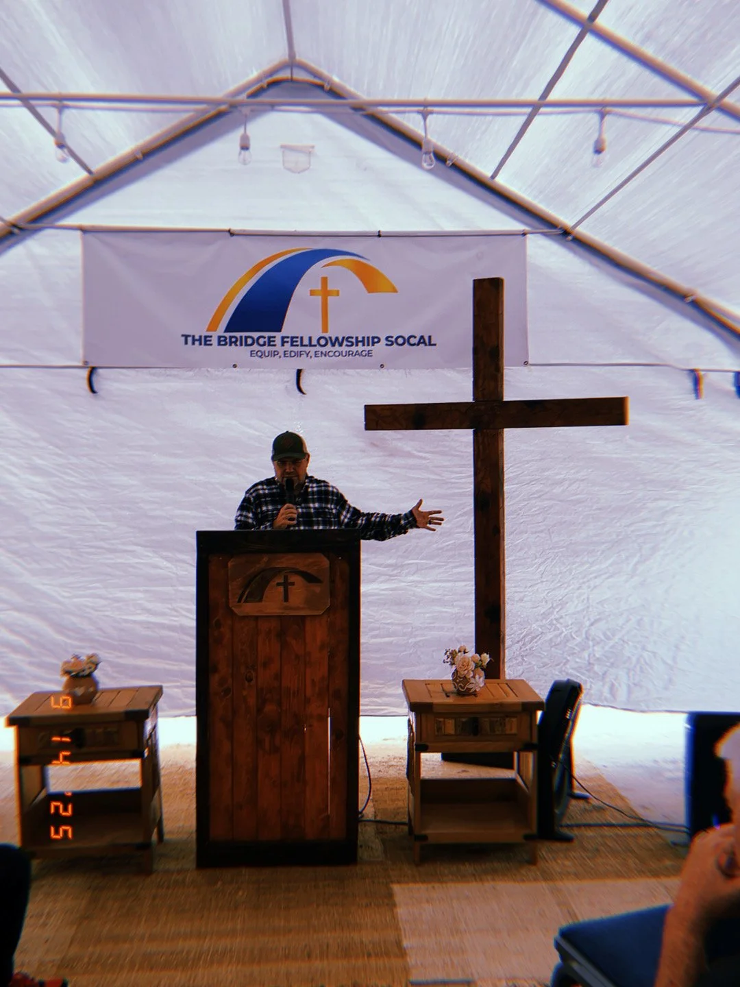 A man standing at a wooden podium with a cross symbol, speaking into a microphone, in a large white tent. Behind him is a large wooden cross and a banner that reads 'The Bridge Fellowship SoCal' with a cross logo. There are small tables with flowers 