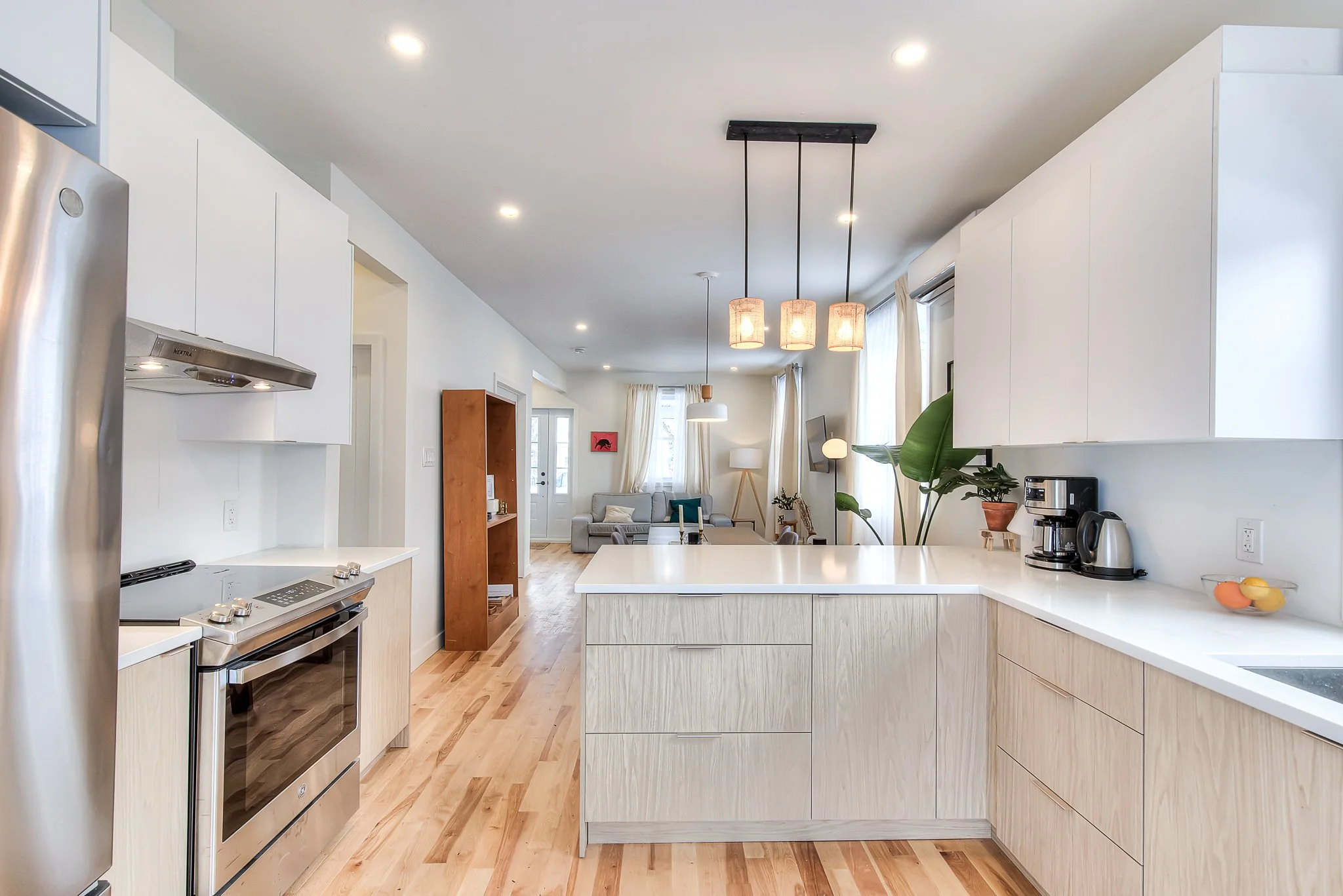 Modern kitchen with white cabinets, stainless steel oven and refrigerator, wooden flooring, and a view into the living room with sofas, plants, and large windows.