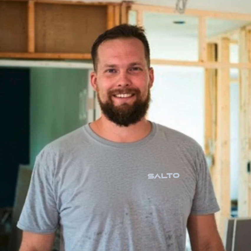 A man with a beard and short hair smiling at a construction site, wearing a gray t-shirt with a logo.