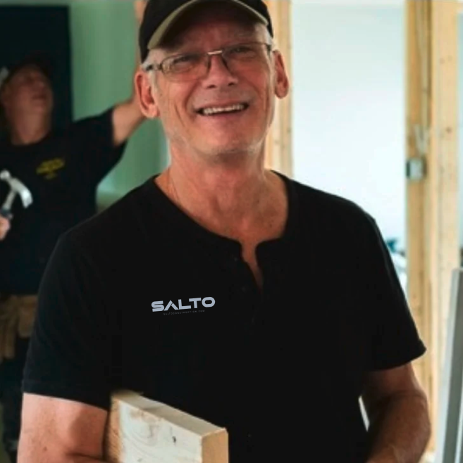 Smiling man wearing a black shirt, glasses, and a cap, holding a piece of wood at a construction site with a person in the background.