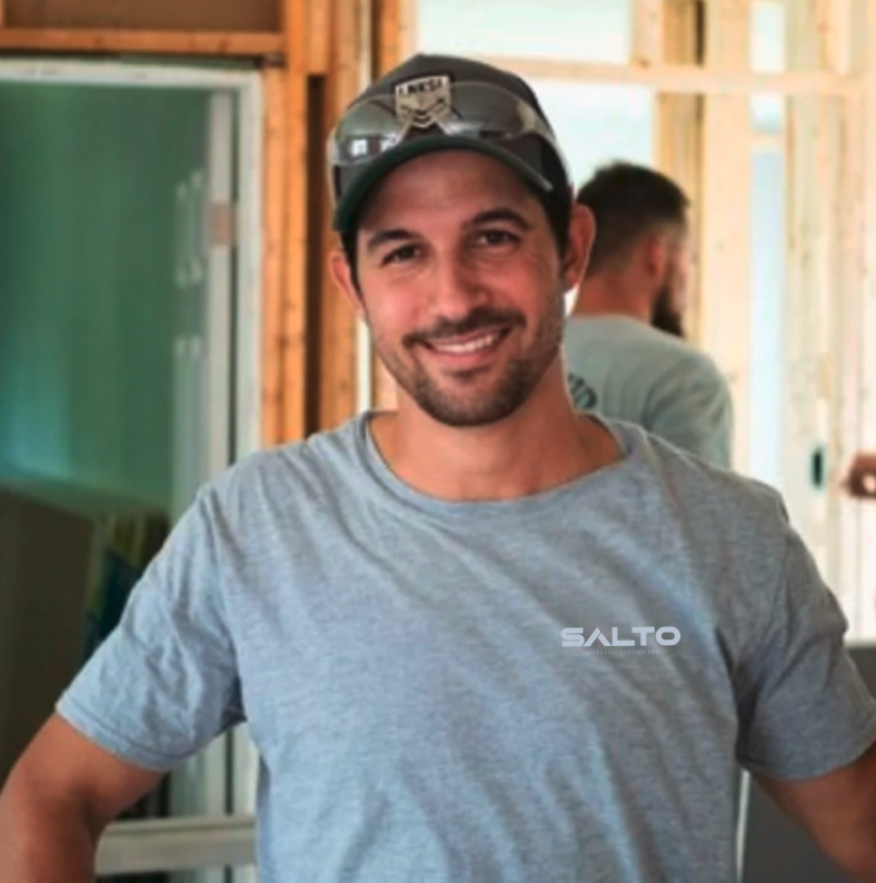 A smiling man wearing a gray t-shirt, sunglasses on his cap, and standing in a construction area with wooden framing behind him.