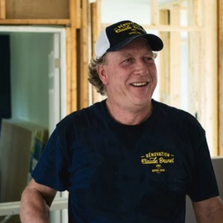 A smiling man wearing a black T-shirt and a black and white trucker hat, standing in a wood-framed construction site.