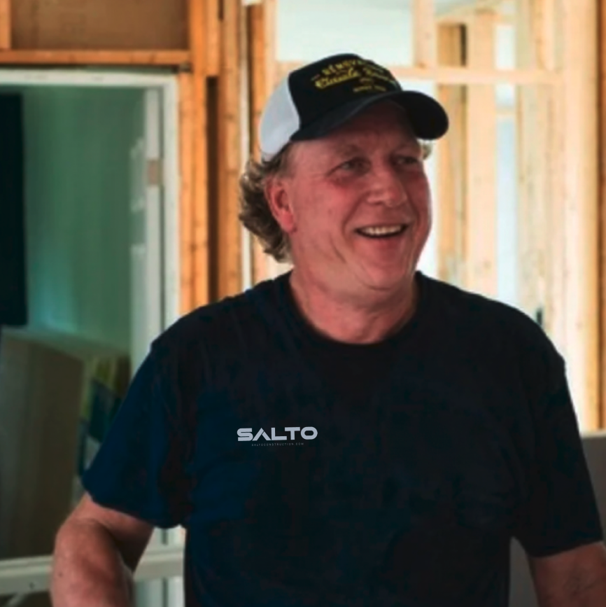 A smiling man wearing a black T-shirt and a black and white trucker hat, standing in a wood-framed construction site.