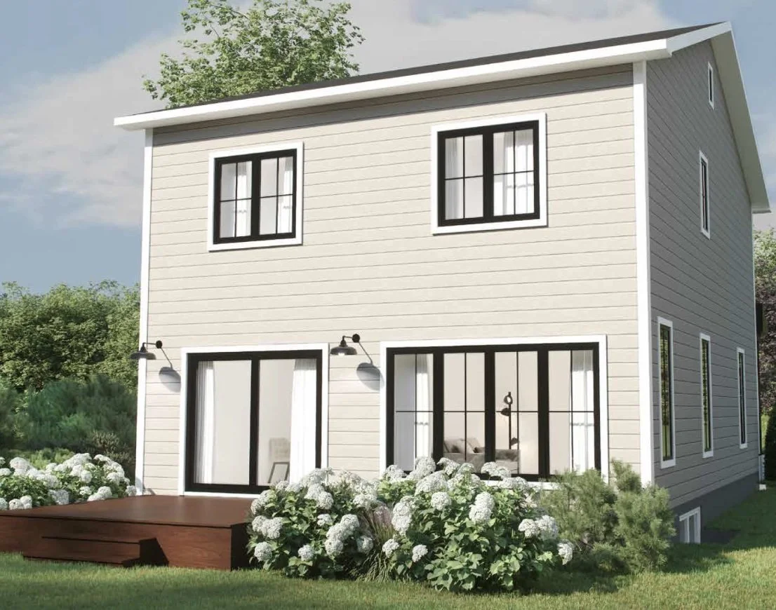 A modern three-story house with beige siding, black-framed windows, and a small wooden deck with white flowering bushes in front, under a partly cloudy sky.