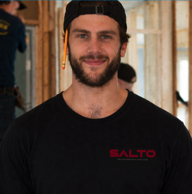 A man with a beard and mustache wearing a black baseball cap backwards and a black t-shirt with a partially visible logo, standing inside a building under construction.