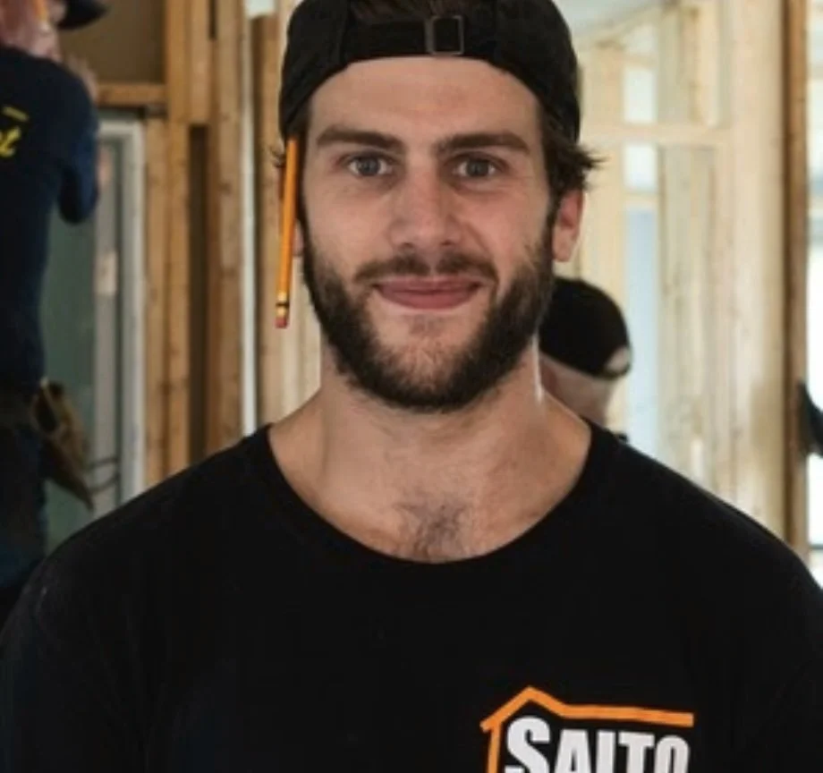 A man with a beard and mustache wearing a black baseball cap backwards and a black t-shirt with a partially visible logo, standing inside a building under construction.