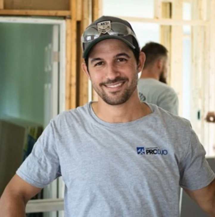 A smiling man wearing a gray t-shirt, sunglasses on his cap, and standing in a construction area with wooden framing behind him.