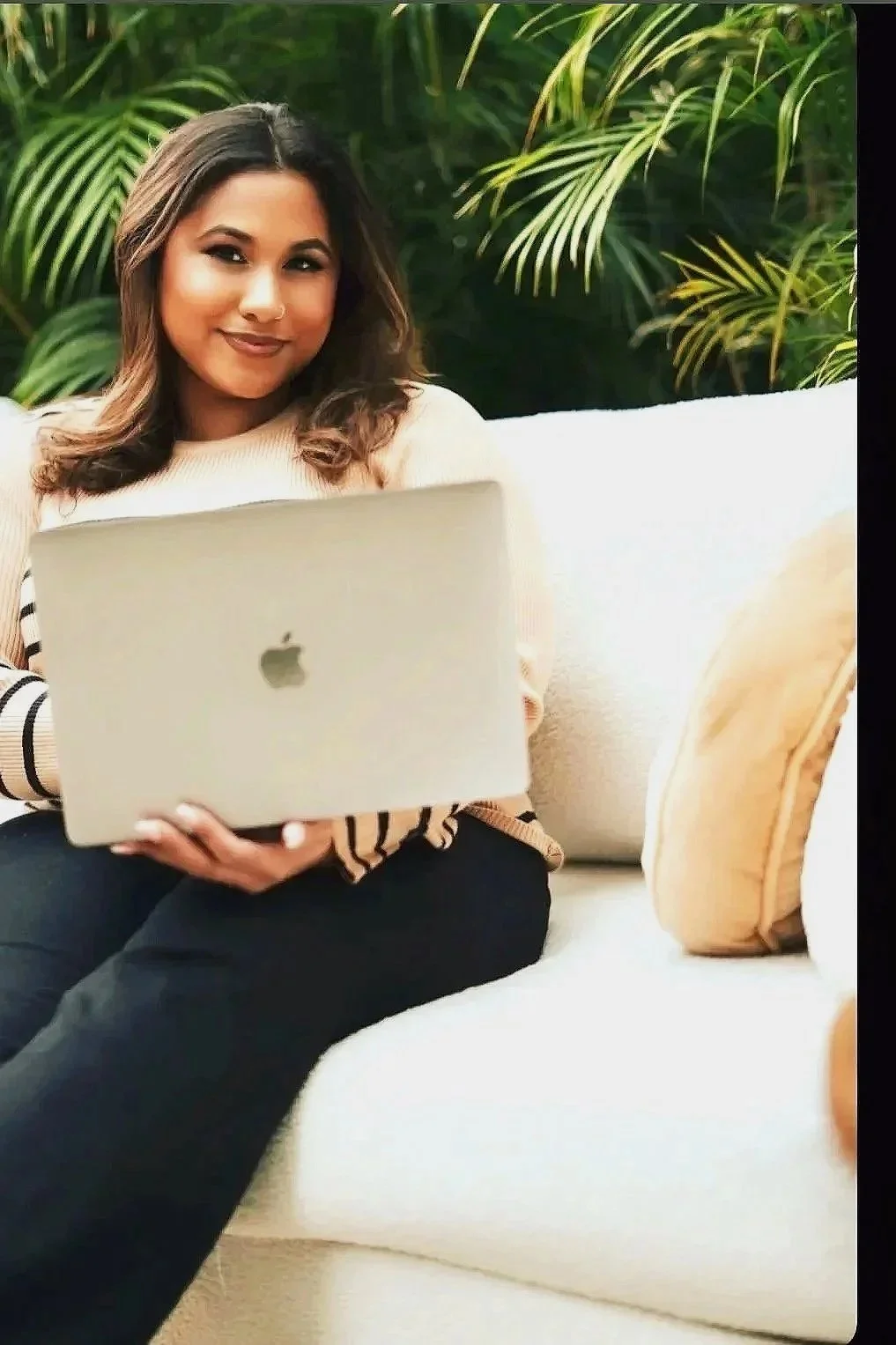 A woman sitting on a white couch holding an open silver MacBook with an apple logo, surrounded by green plants and a beige pillow.