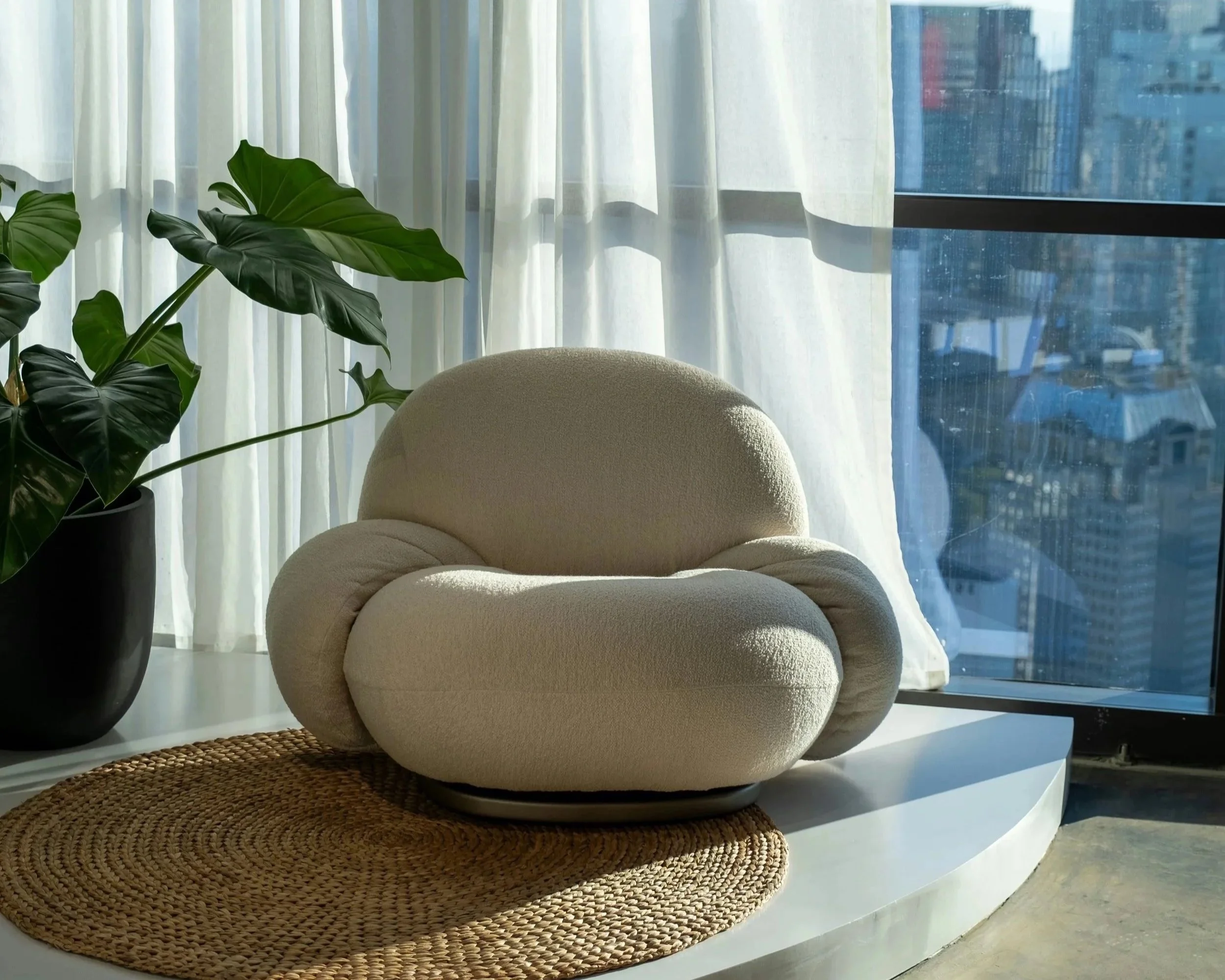 A cream-colored armchair on a round woven rug next to a large potted plant, with sheer curtains and a cityscape view through floor-to-ceiling windows in the background.