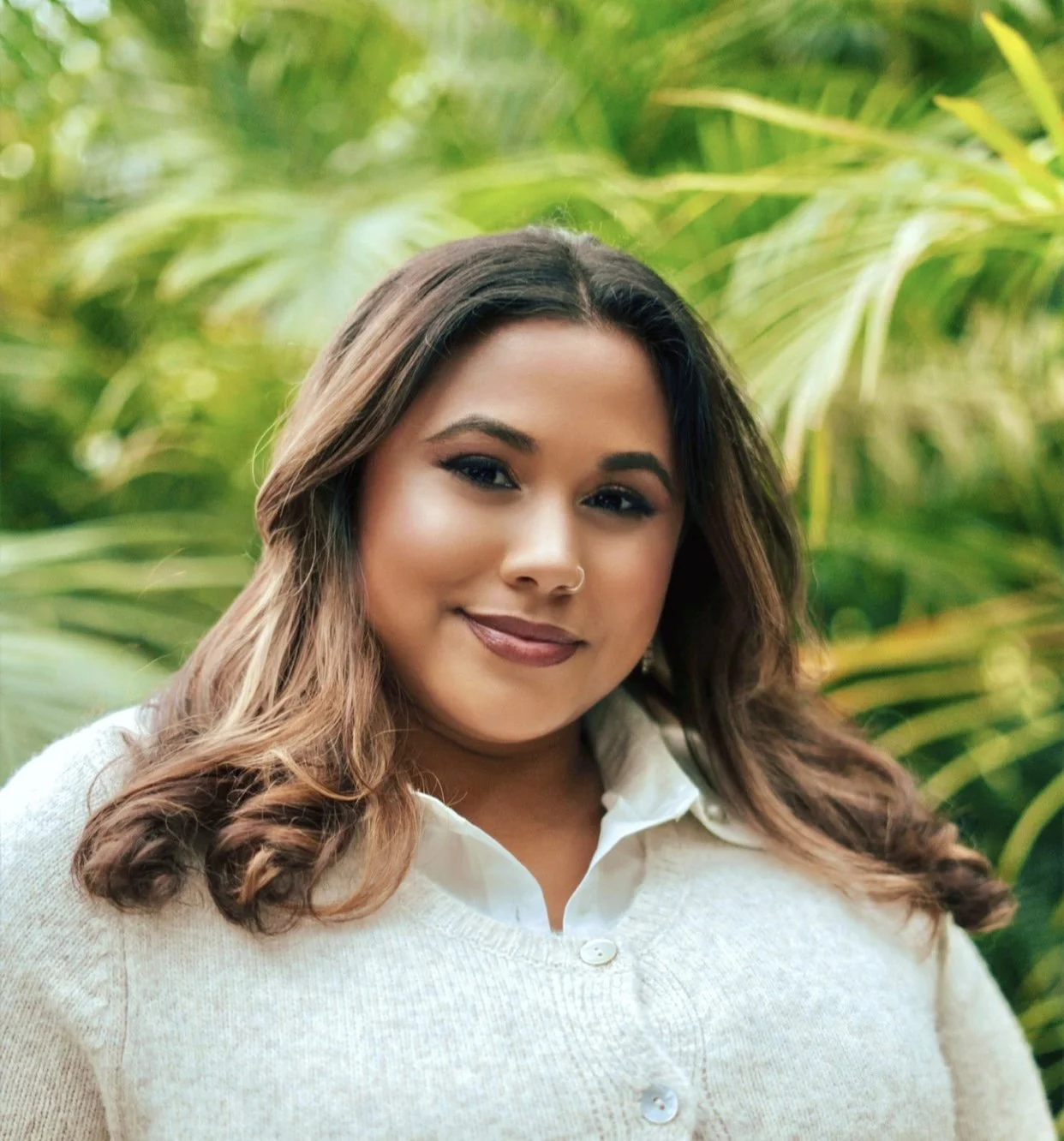 A woman with long, wavy hair and medium brown skin, wearing a white collared shirt and a beige sweater, standing outdoors in front of green foliage, smiling softly.