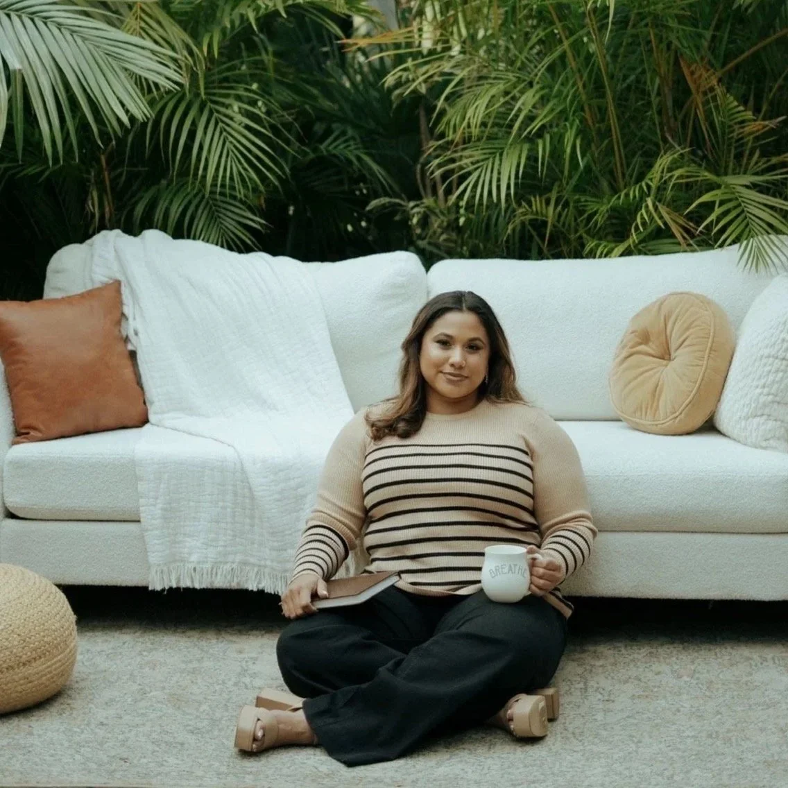A woman sitting cross-legged on a rug in front of a white sofa with multiple pillows, holding a mug and a notebook, with large green plants in the background.